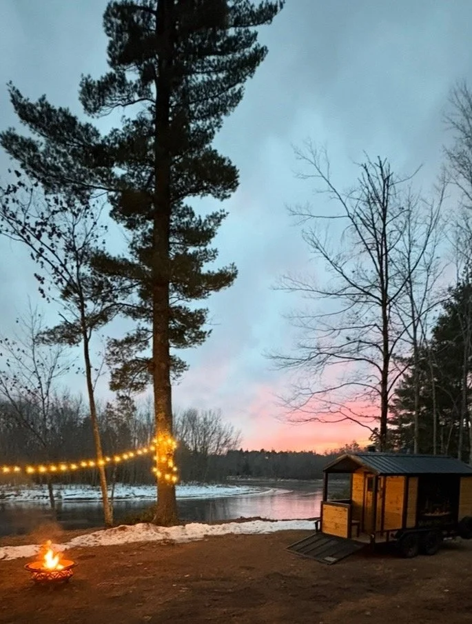 A lakeside scene at sunset with a tall pine tree wrapped in string lights, a small fire pit on the ground, and a wooden trailer on wheels. Bare trees are visible around the lake, and the sky has pink and blue hues.
