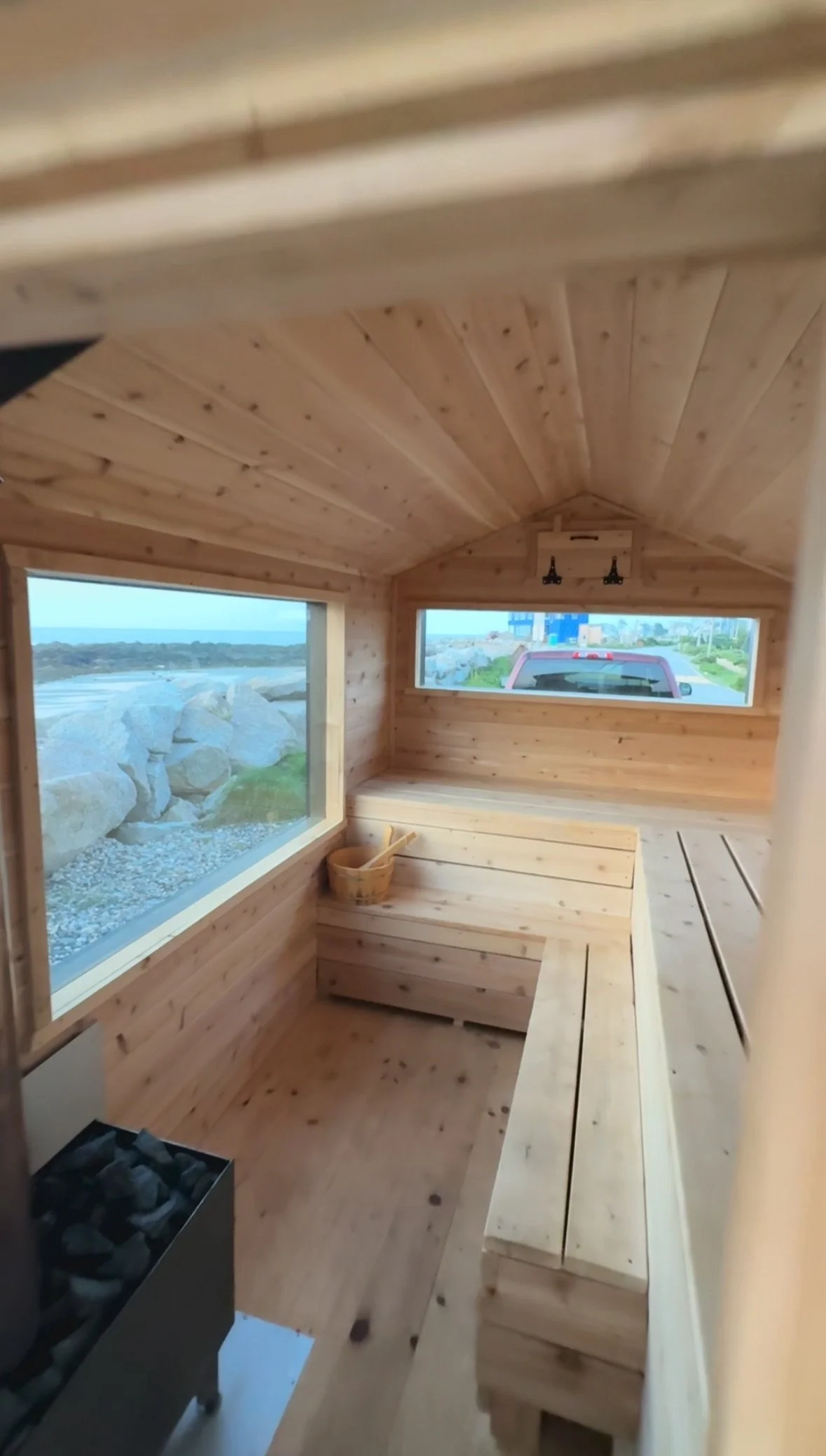 Interior of a small wooden sauna with large windows showing outdoor landscape and a red truck, featuring wooden benches and a bucket with a ladle inside.