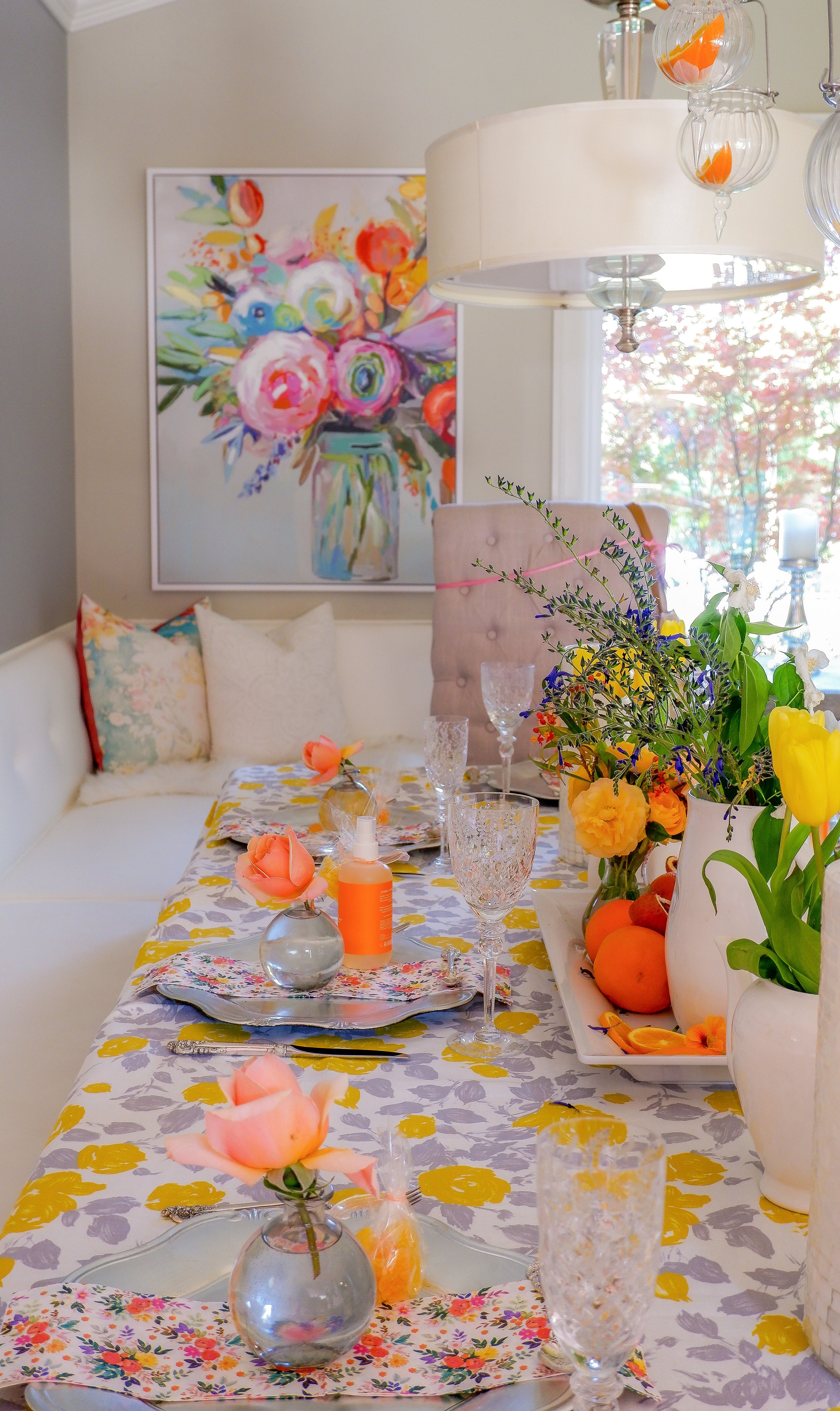 A brightly decorated dining table with floral tablecloth, floral plates, and crystal glasses. Pink and orange flowers in small vases and a white vase with yellow and purple flowers are on the table. Nearby, there are oranges and a vibrant painting of flowers on the wall.