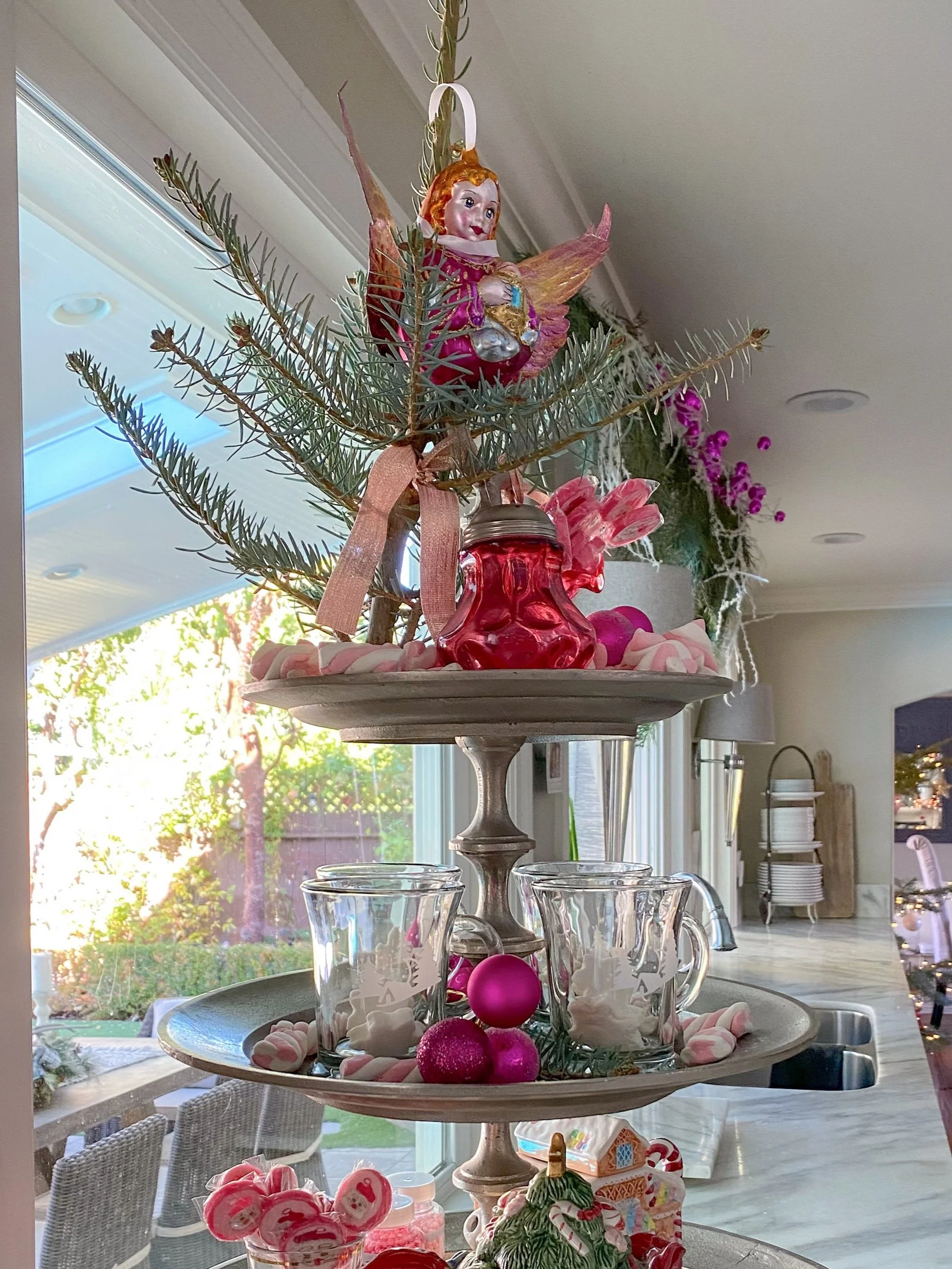 A three-tiered tray with holiday decorations, including a small angel figurine, pink and red ornaments, and festive mugs with reindeer designs. It is adorned with sprigs of evergreen and surrounded by pink candies. The setting is a kitchen with a window view in the background.