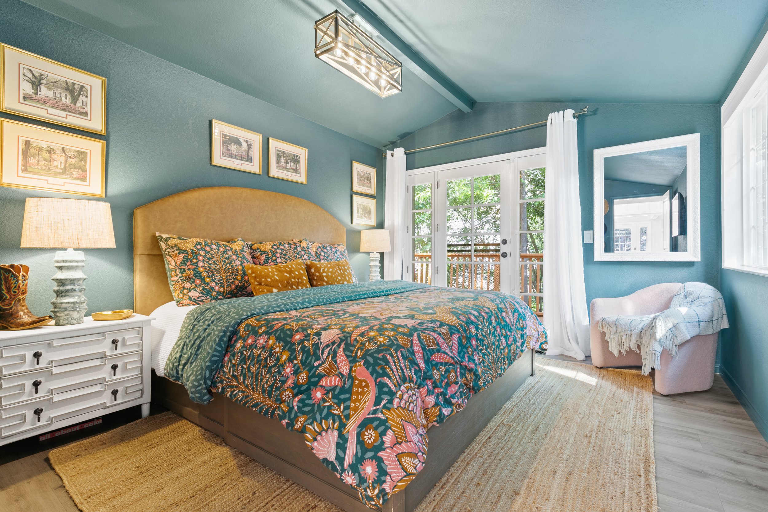 Cozy bedroom with teal walls, floral bedding, and white curtains. Features a wicker rug, a mirrored wall, framed art, and a decorative chair with a throw blanket.