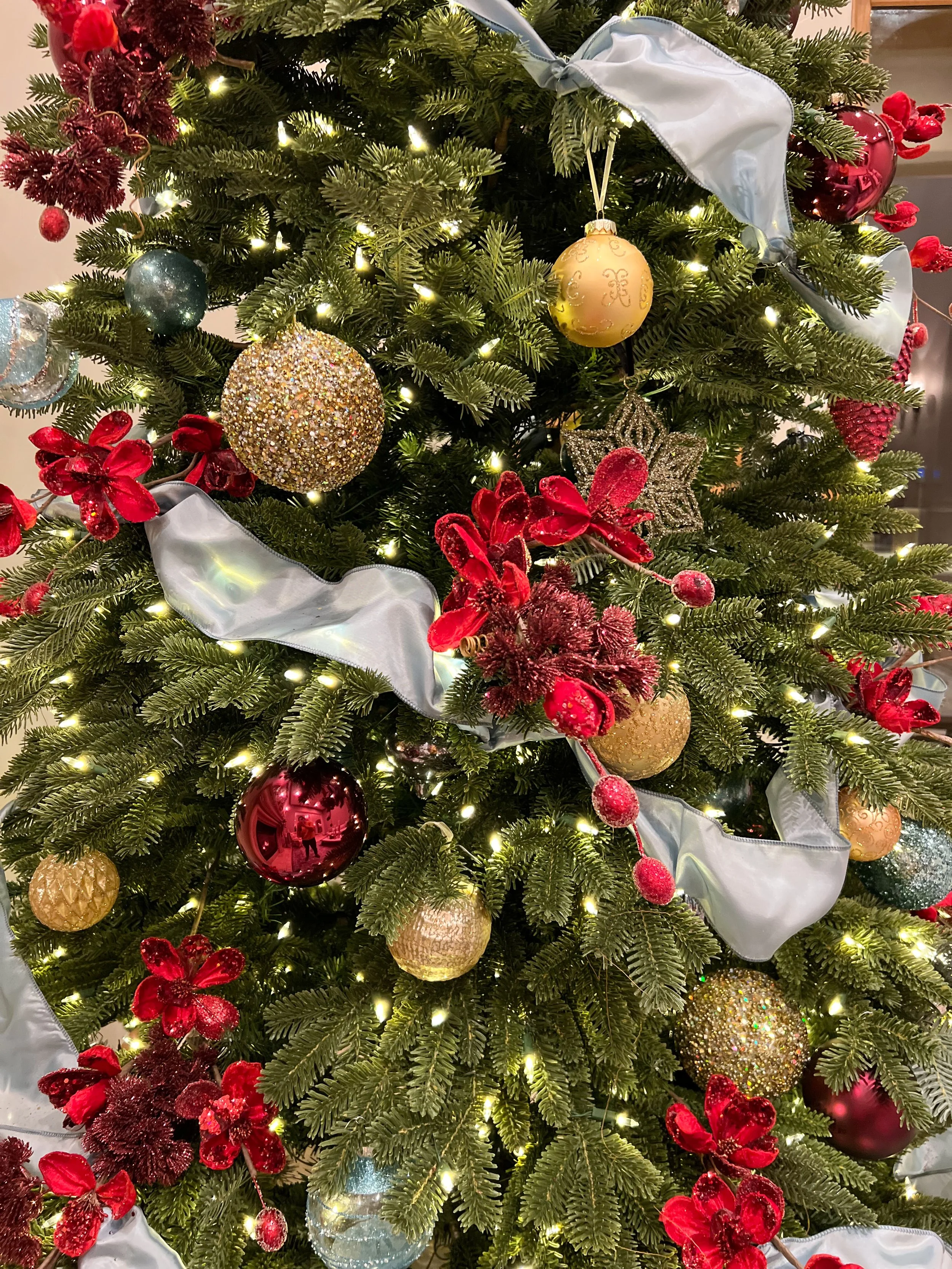Decorated Christmas tree with red flowers, gold, silver, and red ornaments, and white ribbon.