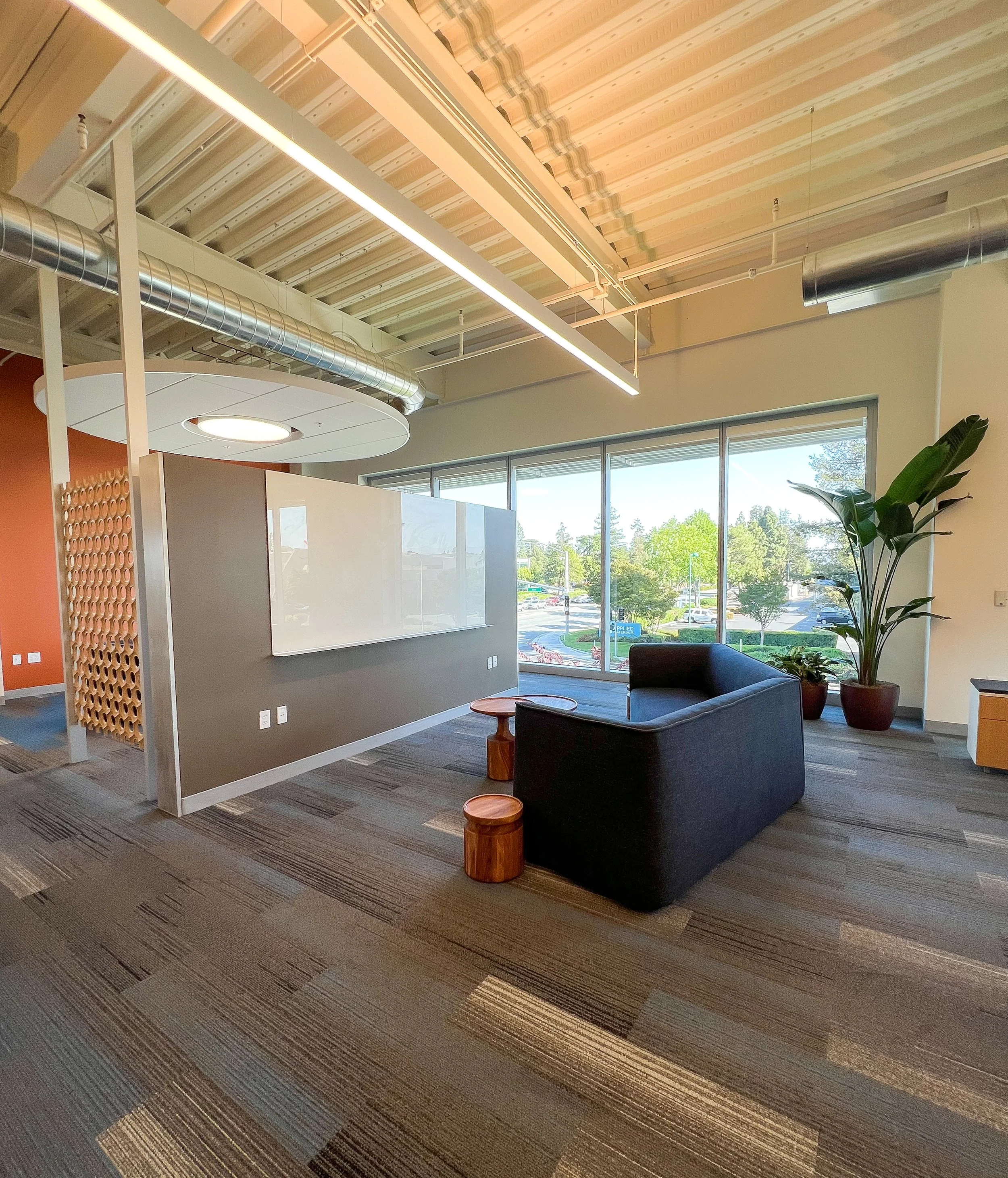 Modern office space with large windows, a black sofa, wooden stools, a potted plant, and a whiteboard on a partition wall. Exposed ceiling with ductwork is visible.