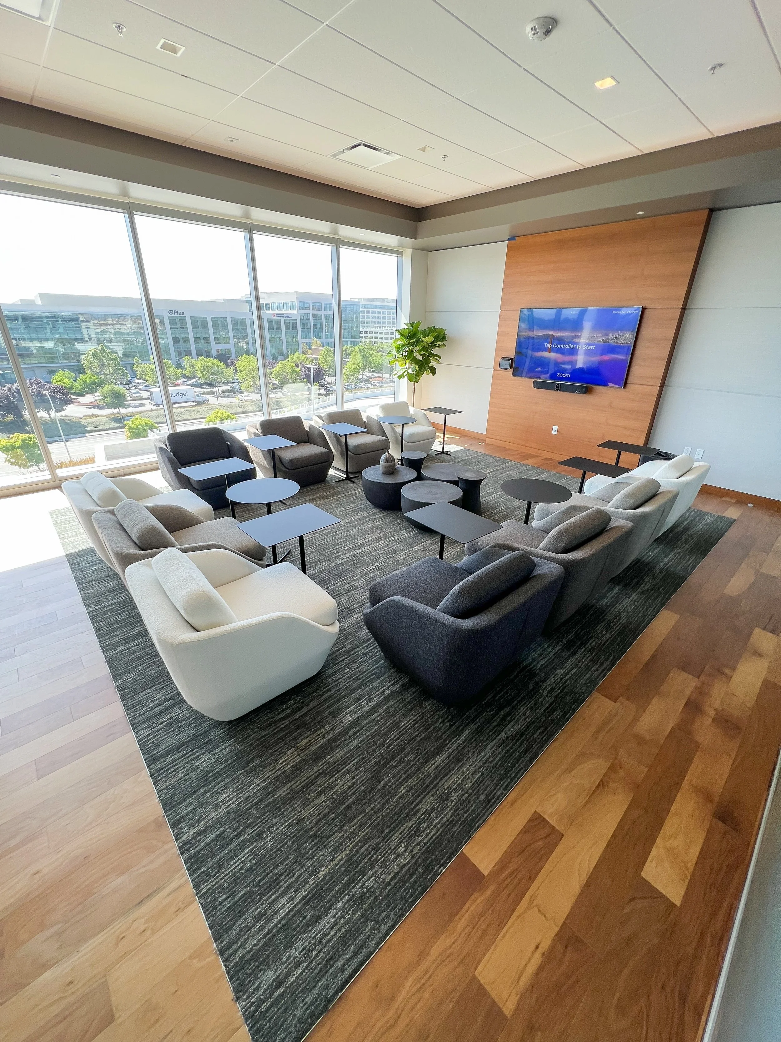 Modern office lounge with armchairs, tables, wall-mounted TV and large windows.