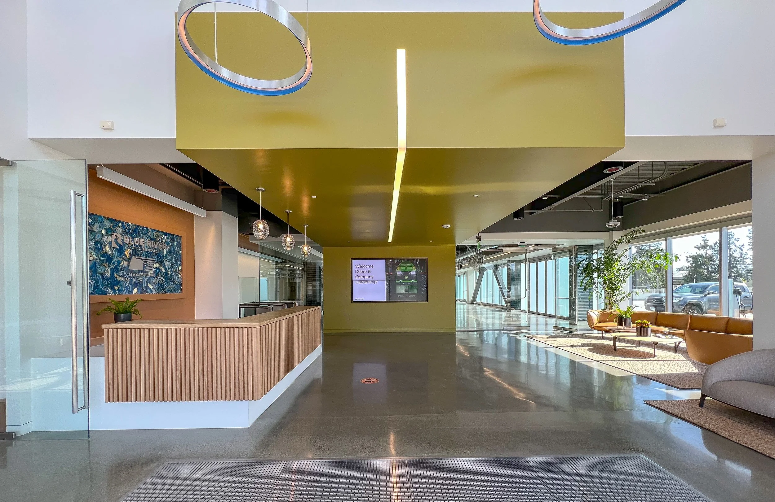 Modern office lobby with wooden reception desk, decorative lights, seating area, and large windows.