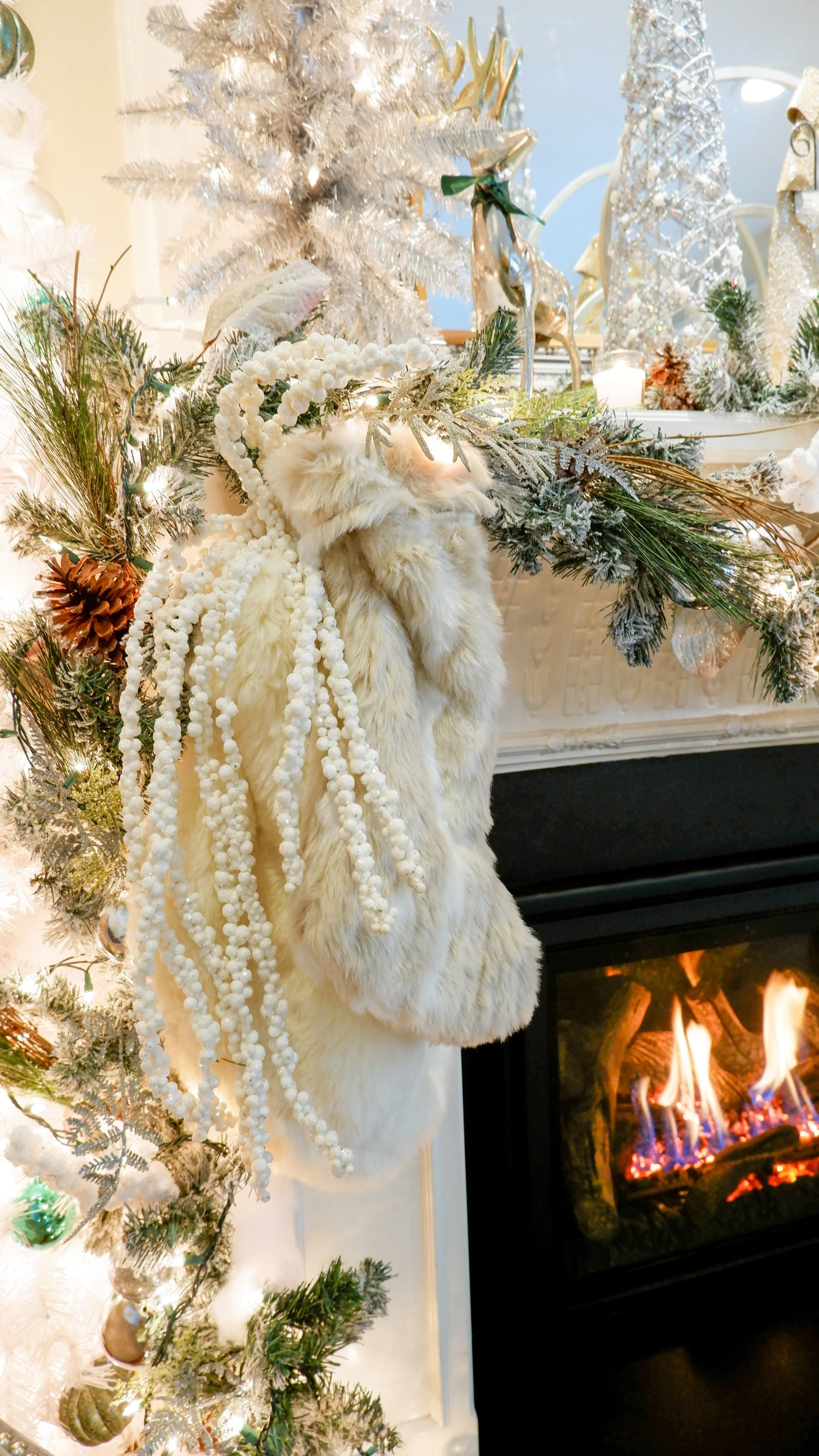 Christmas mantel decorated with fur stocking, pine garland, silver reindeer, and white ornaments next to a glowing fireplace.