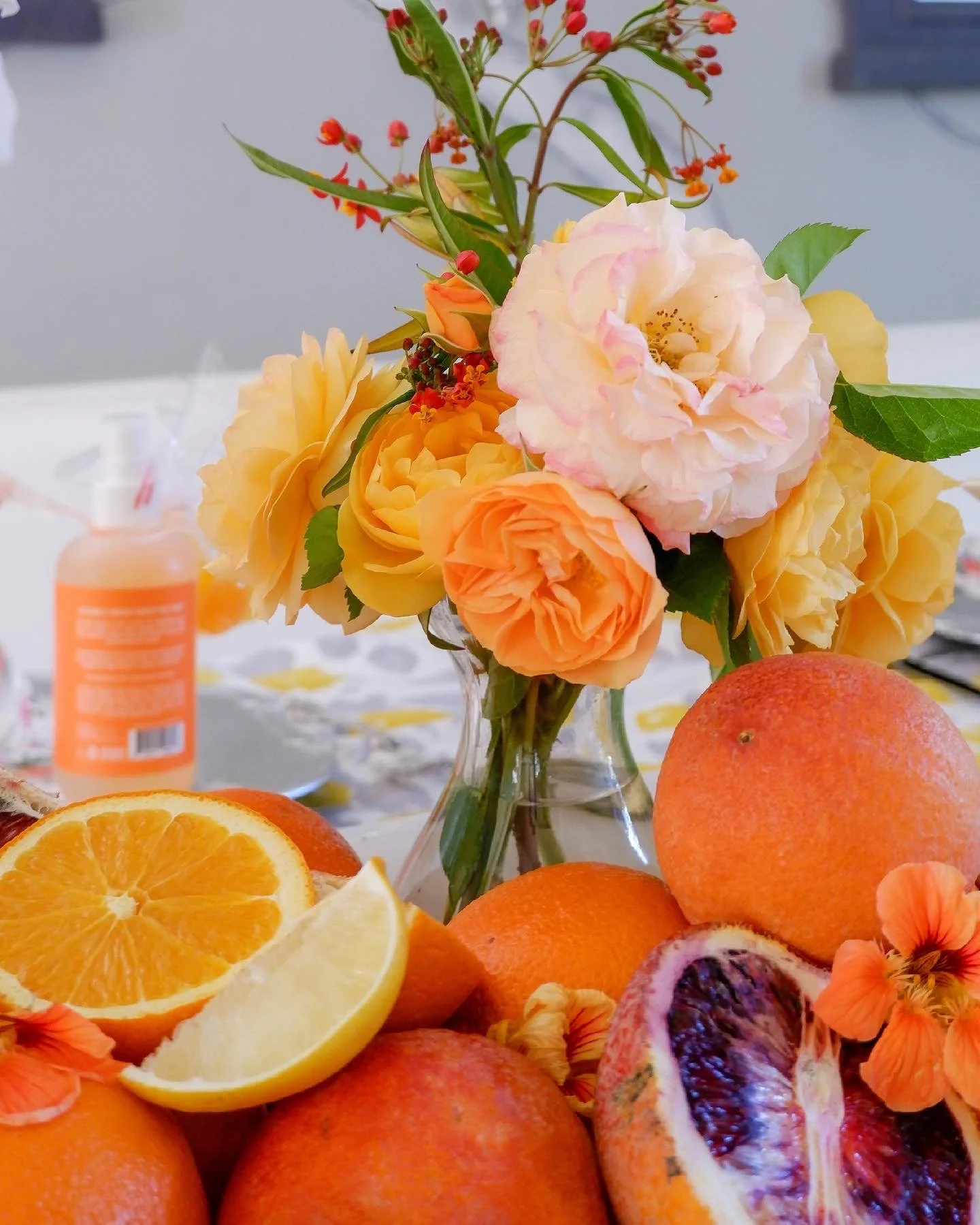 Flowers in vase with oranges, blood oranges, and lemon slices on a table, with a blurry bottle in the background.
