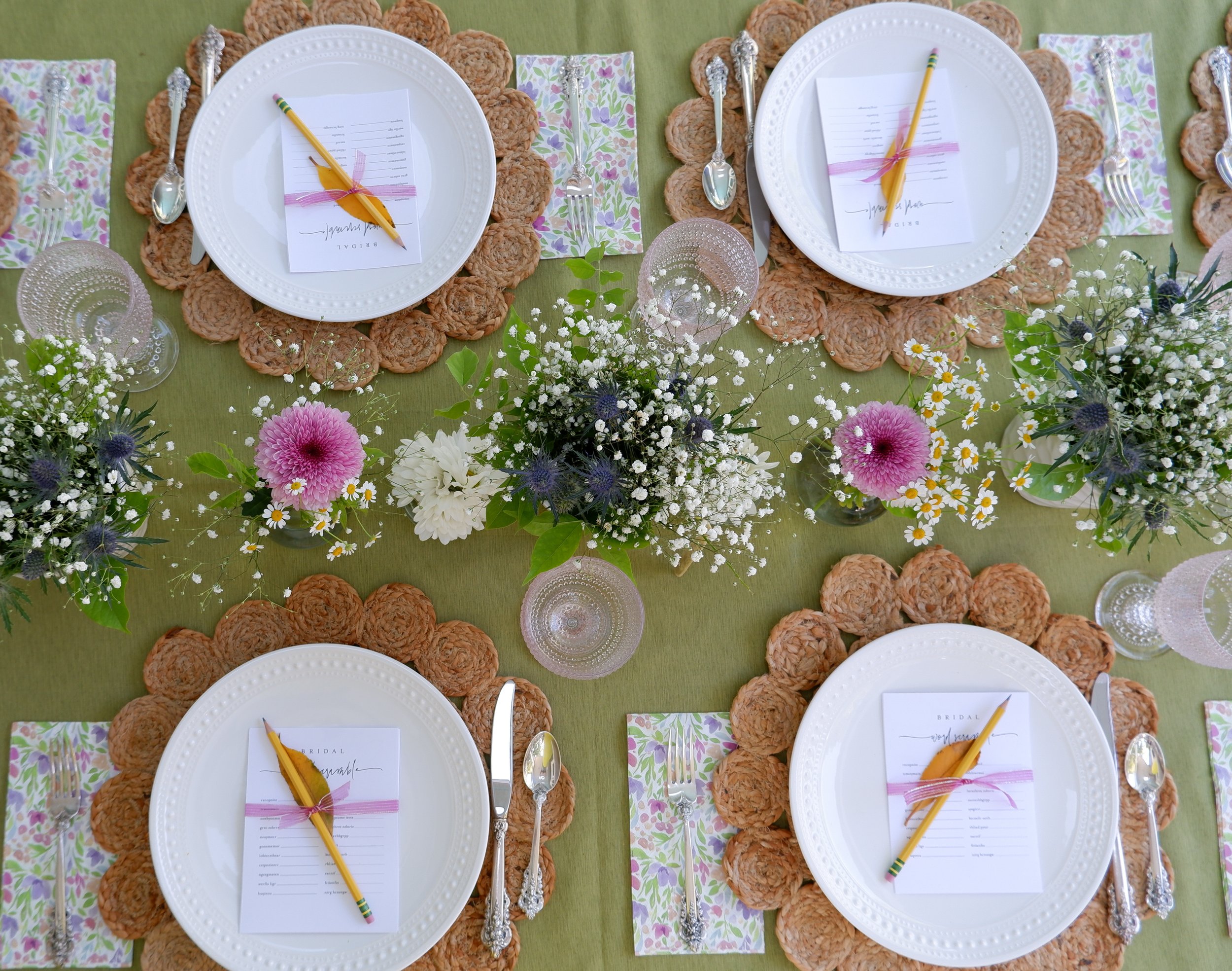Elegant table setting with green tablecloth, white plates on woven placemats, floral napkins, and silver cutlery. Centerpieces feature white, pink, and purple flowers with greenery. Each plate has a menu card and a pencil tied with ribbon.