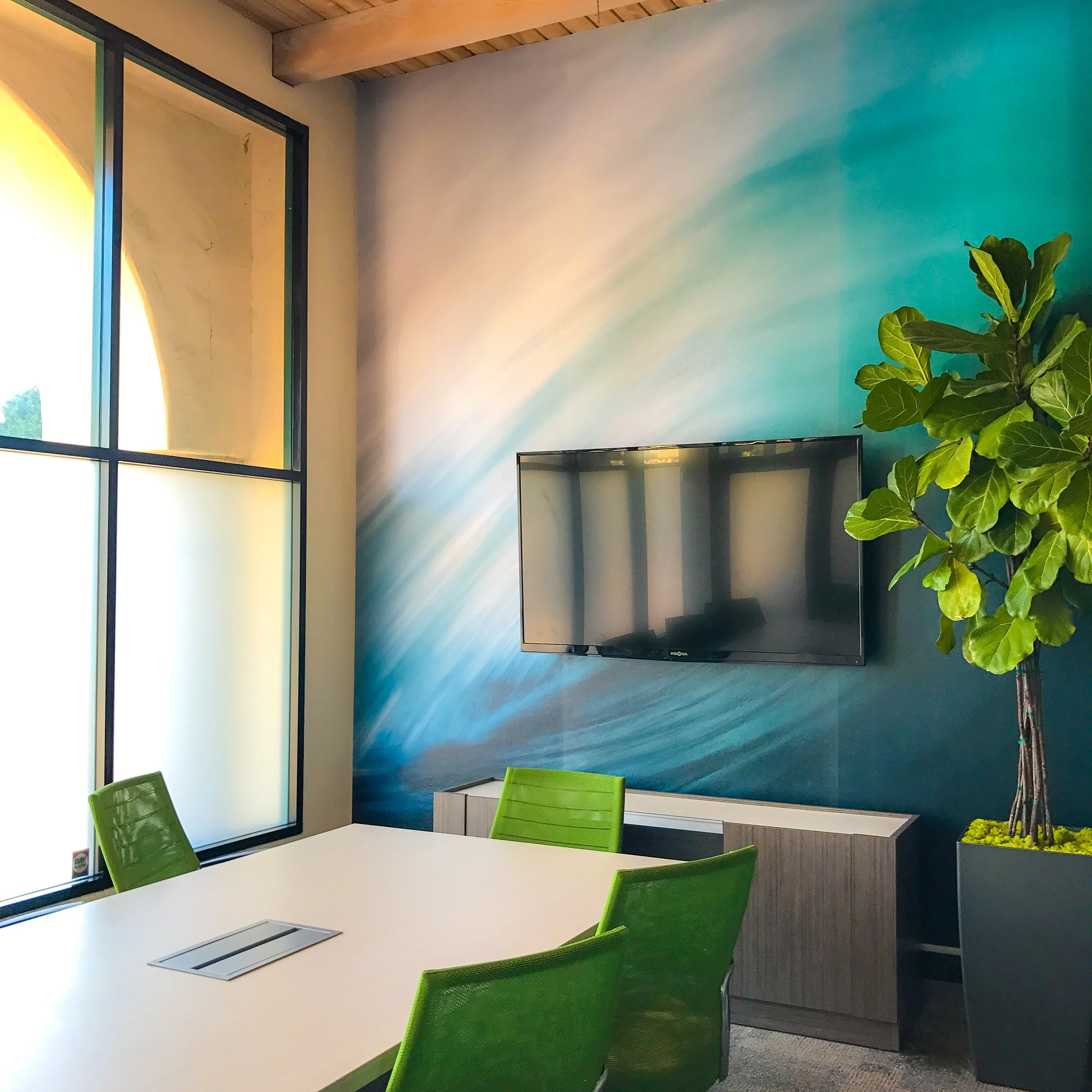 Modern conference room with a table, green chairs, wall-mounted TV, abstract blue wall mural, large plant, and window.