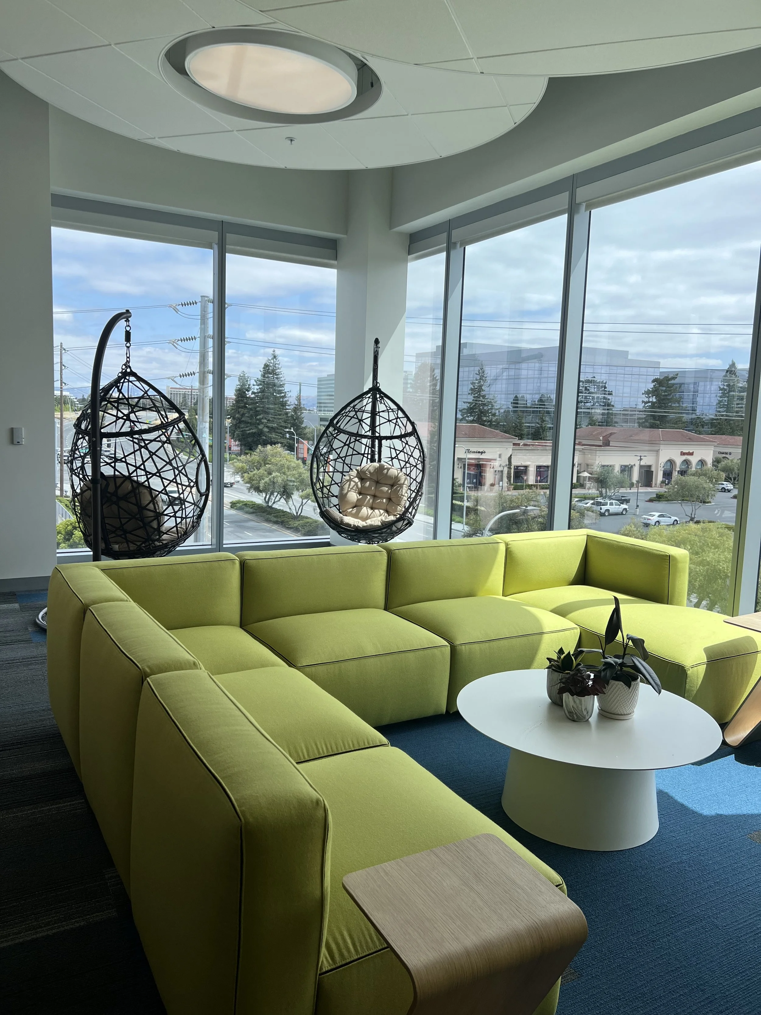 Modern office lounge with lime green sectional sofa, hanging wicker egg chairs, white round coffee table, and large windows with outdoor view.