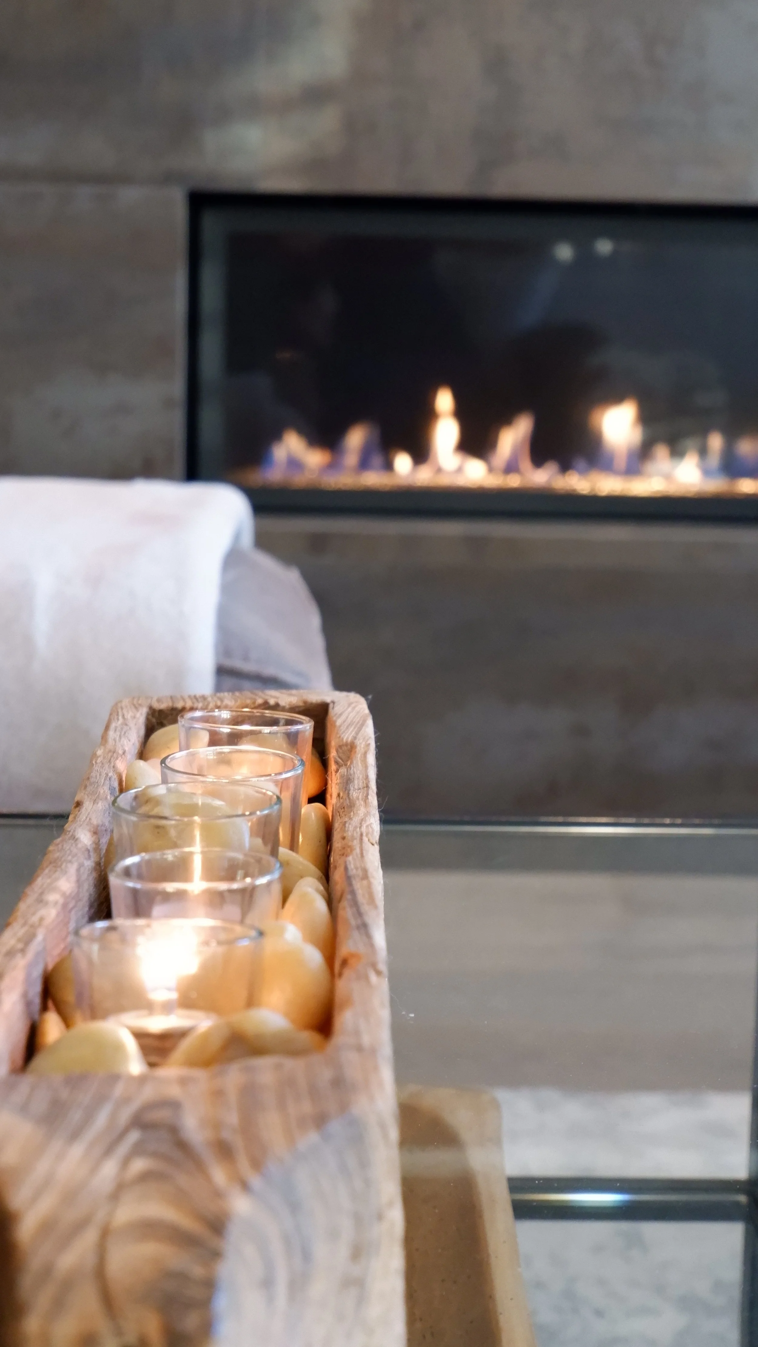 Modern living room with a lit fireplace in the background, and a wooden tray with glass votive candle holders on a glass table in the foreground.