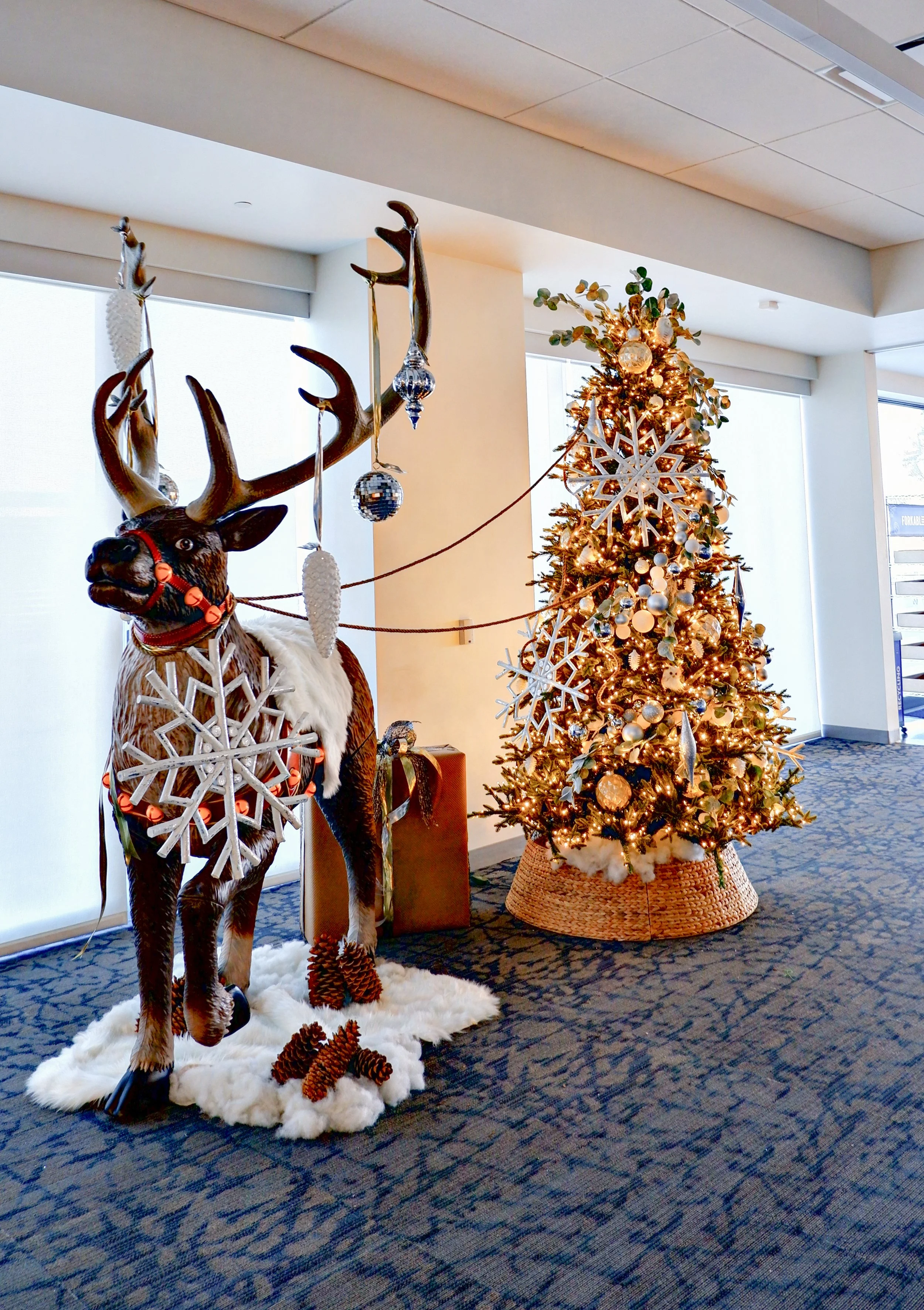 Christmas decoration featuring a large reindeer sculpture with decorative snowflakes, ornaments, and pinecones, standing next to a decorated Christmas tree with lights and ornaments.