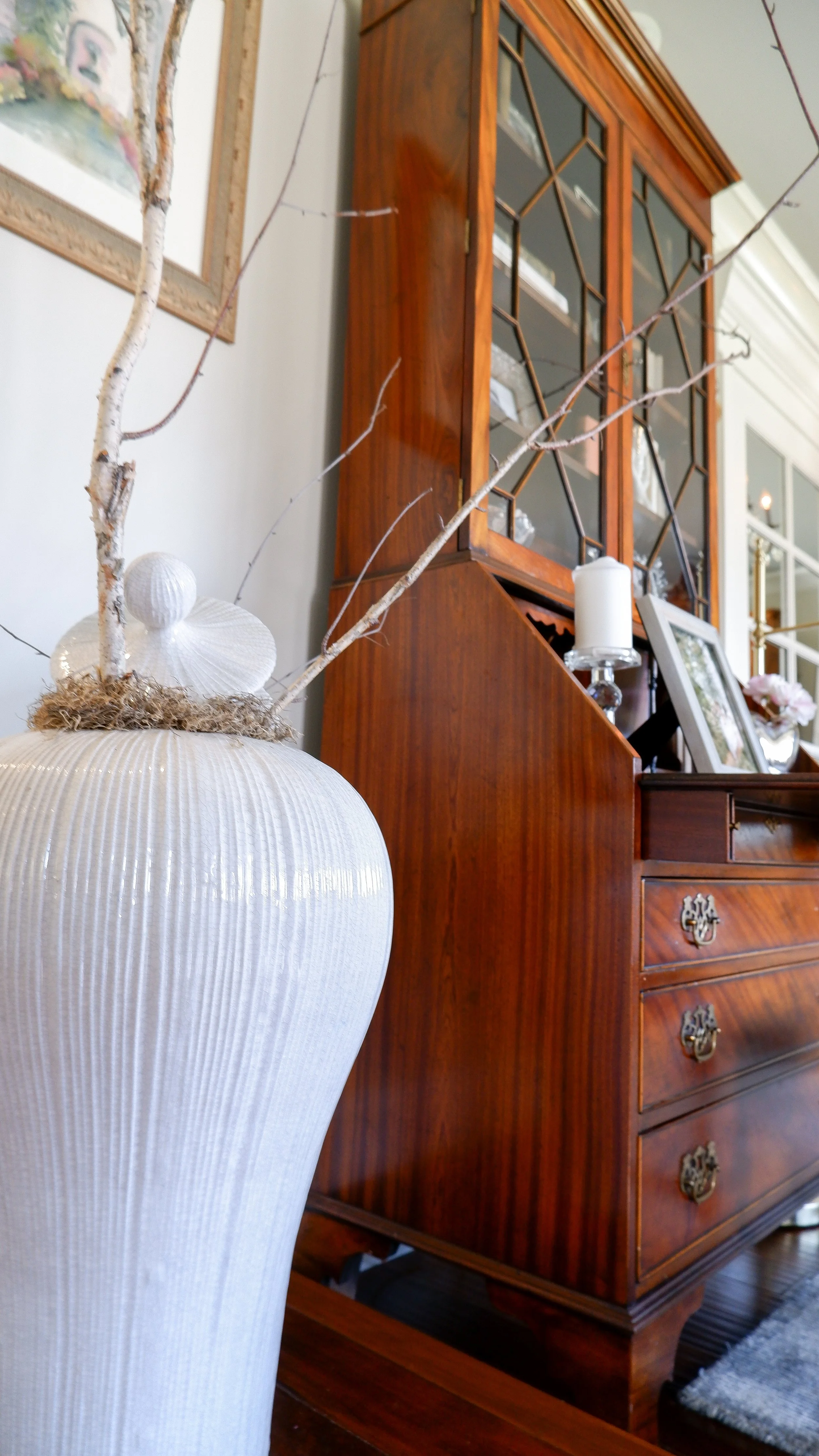 White ceramic vase with twigs placed next to a wooden cabinet in a living room.