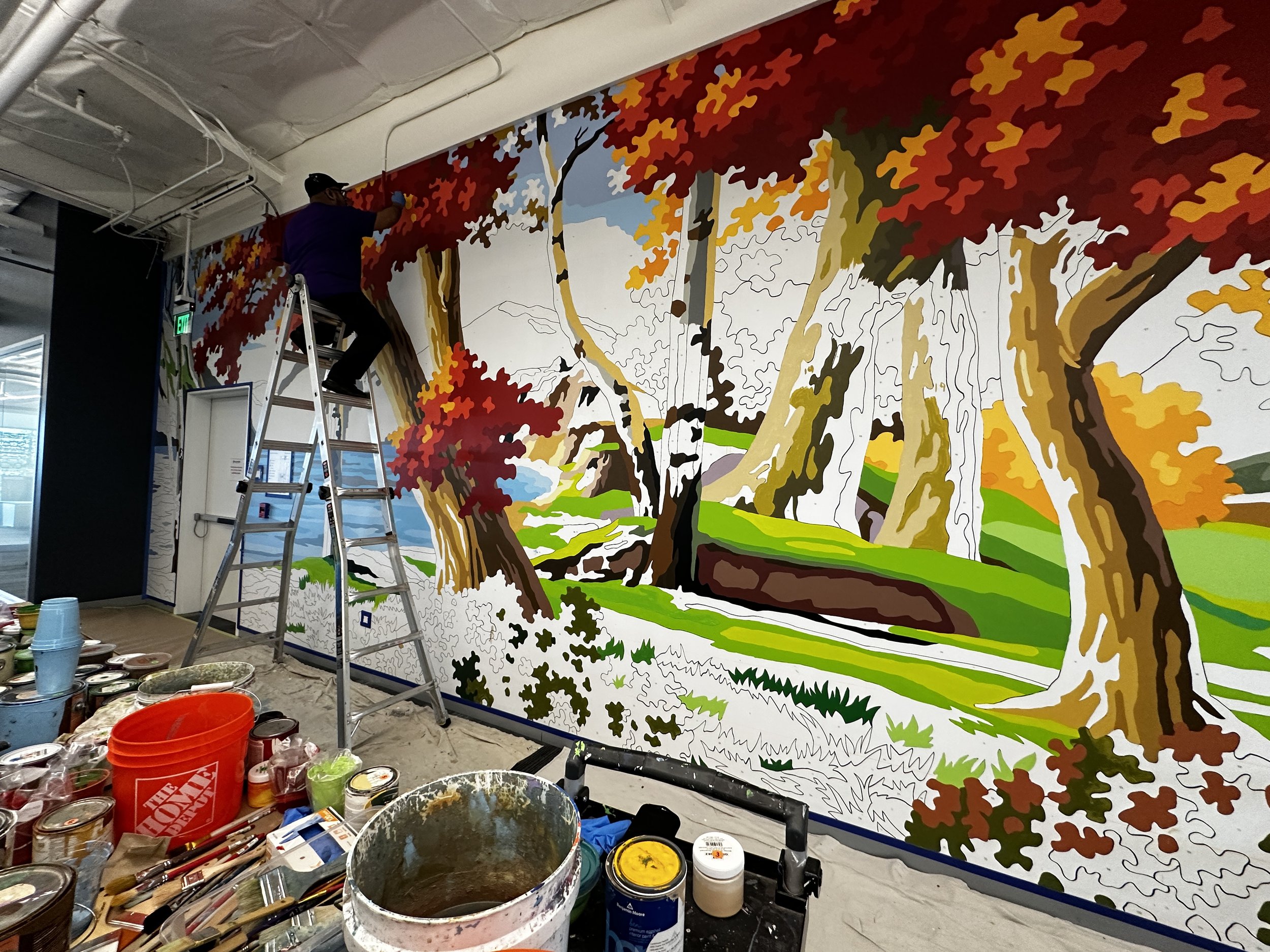 Artist painting a colorful mural of a forest scene with autumn trees on a large indoor wall using a ladder, surrounded by paint cans and brushes.