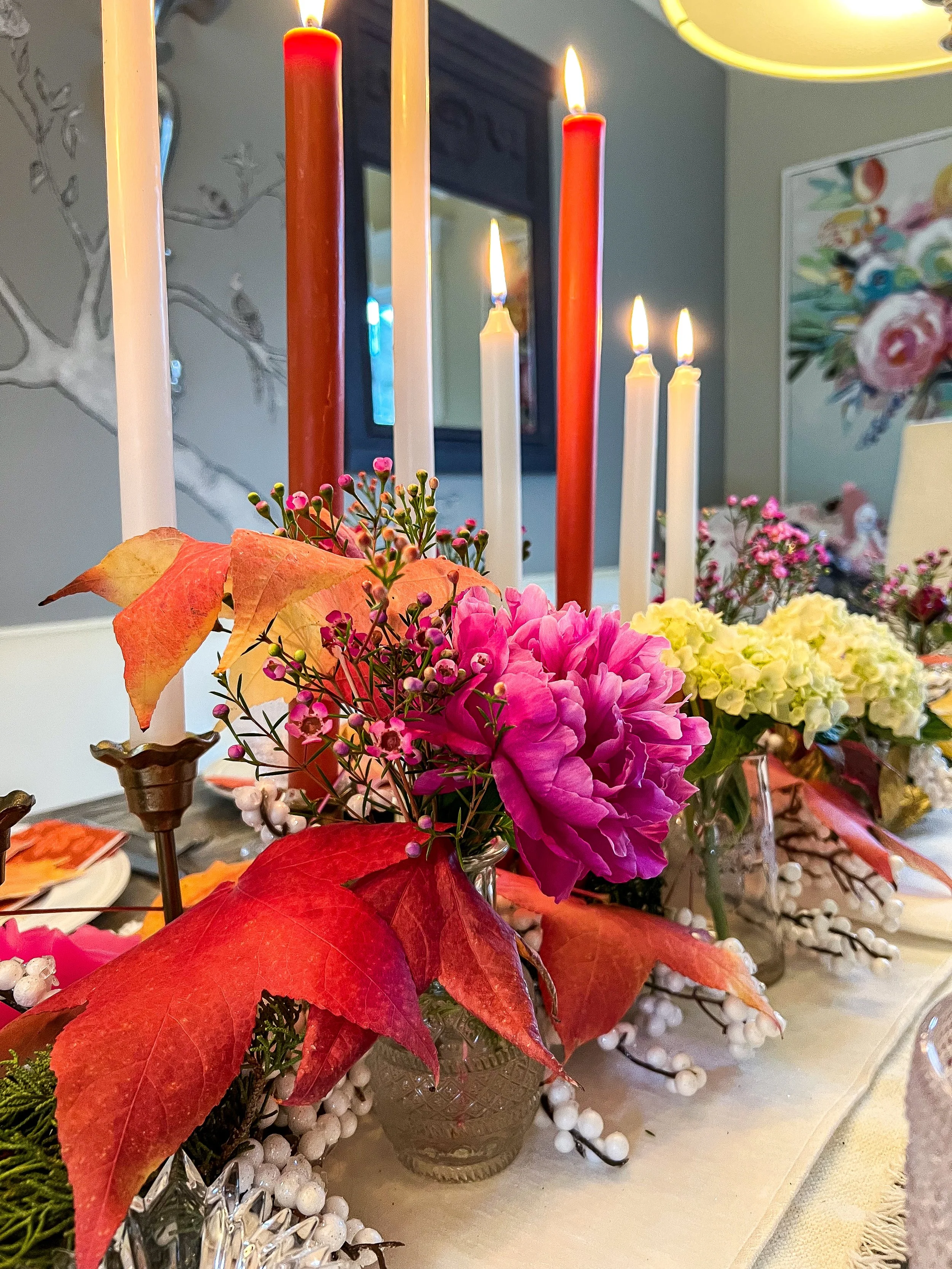 A decorative table setting with red and white candles, vibrant autumn leaves, pink and white flowers, and berry sprigs arranged in glass vases.