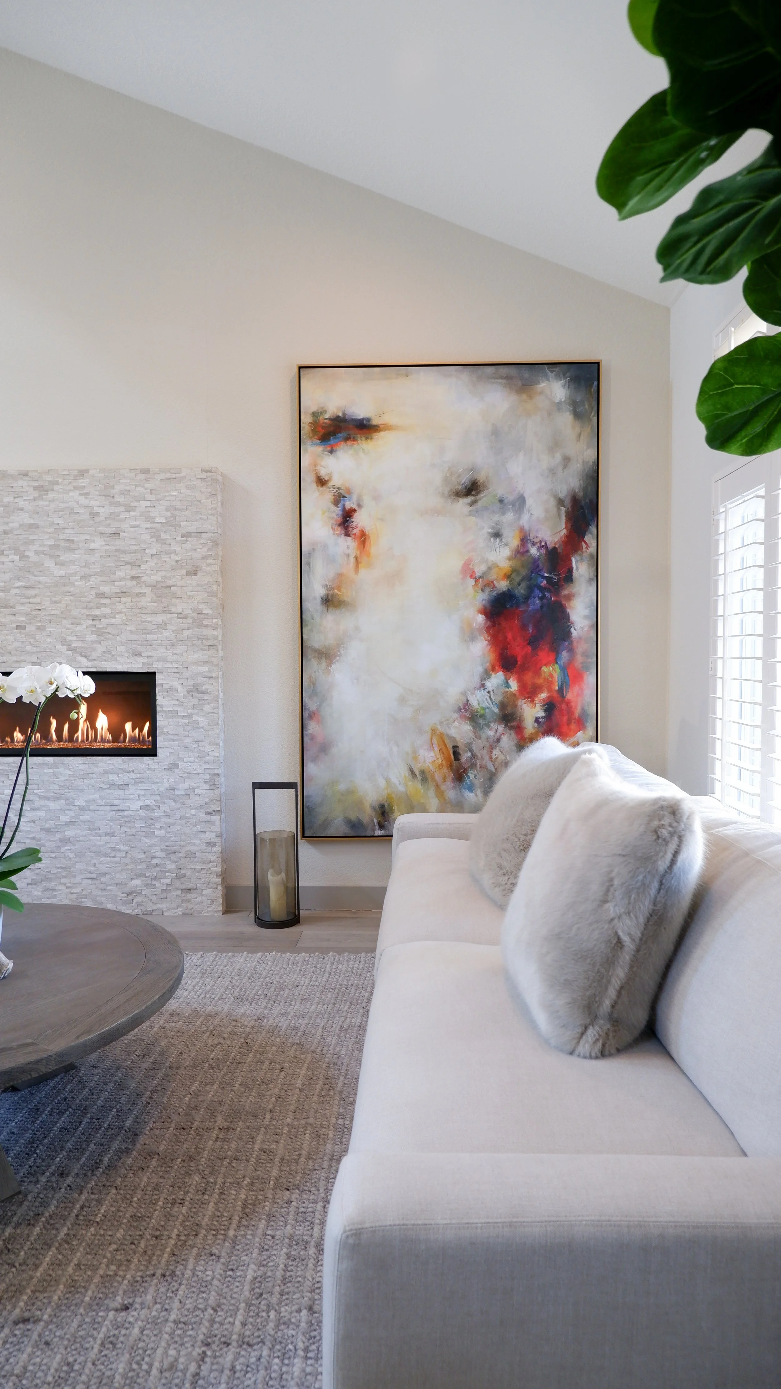 Modern living room with abstract painting, beige sofa, textured rug, stone fireplace, and green plant.