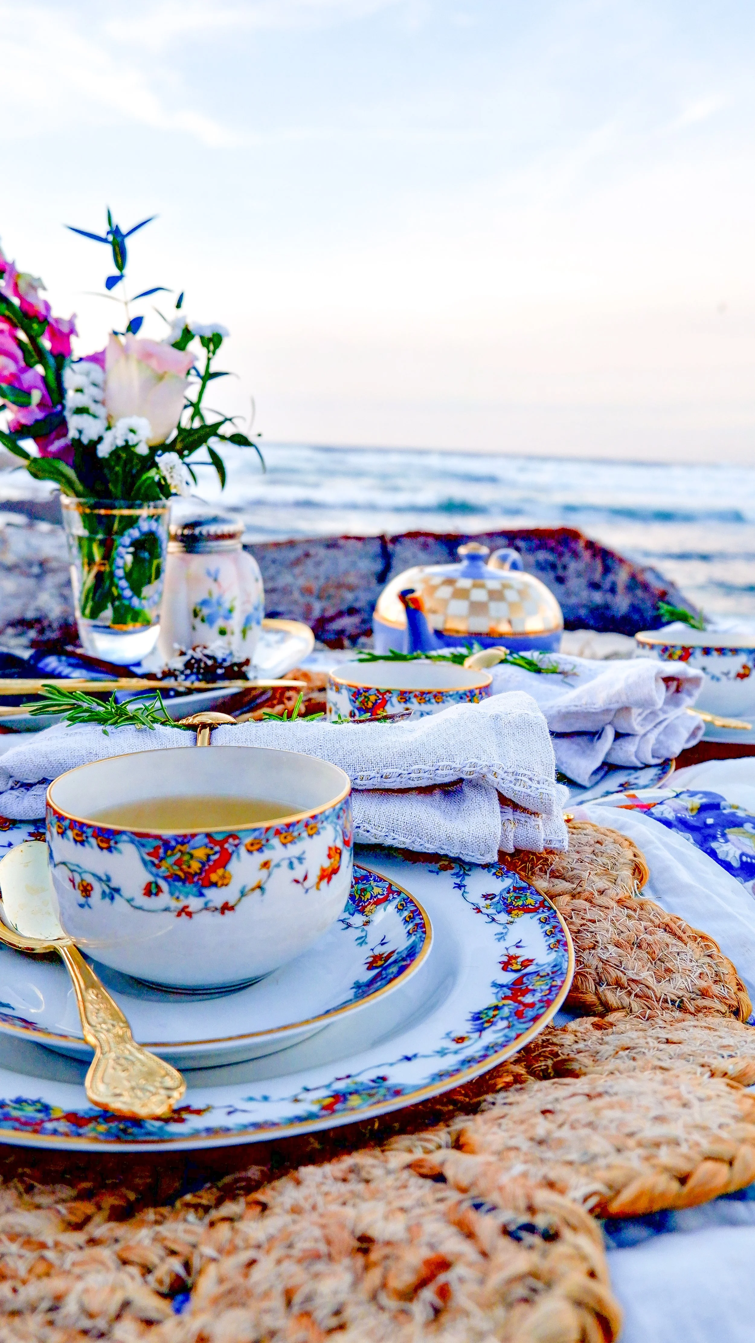 Elegant tea set on a mat by the seaside with floral-patterned cup and saucer, golden spoon, folded napkins, and a small vase with flowers.