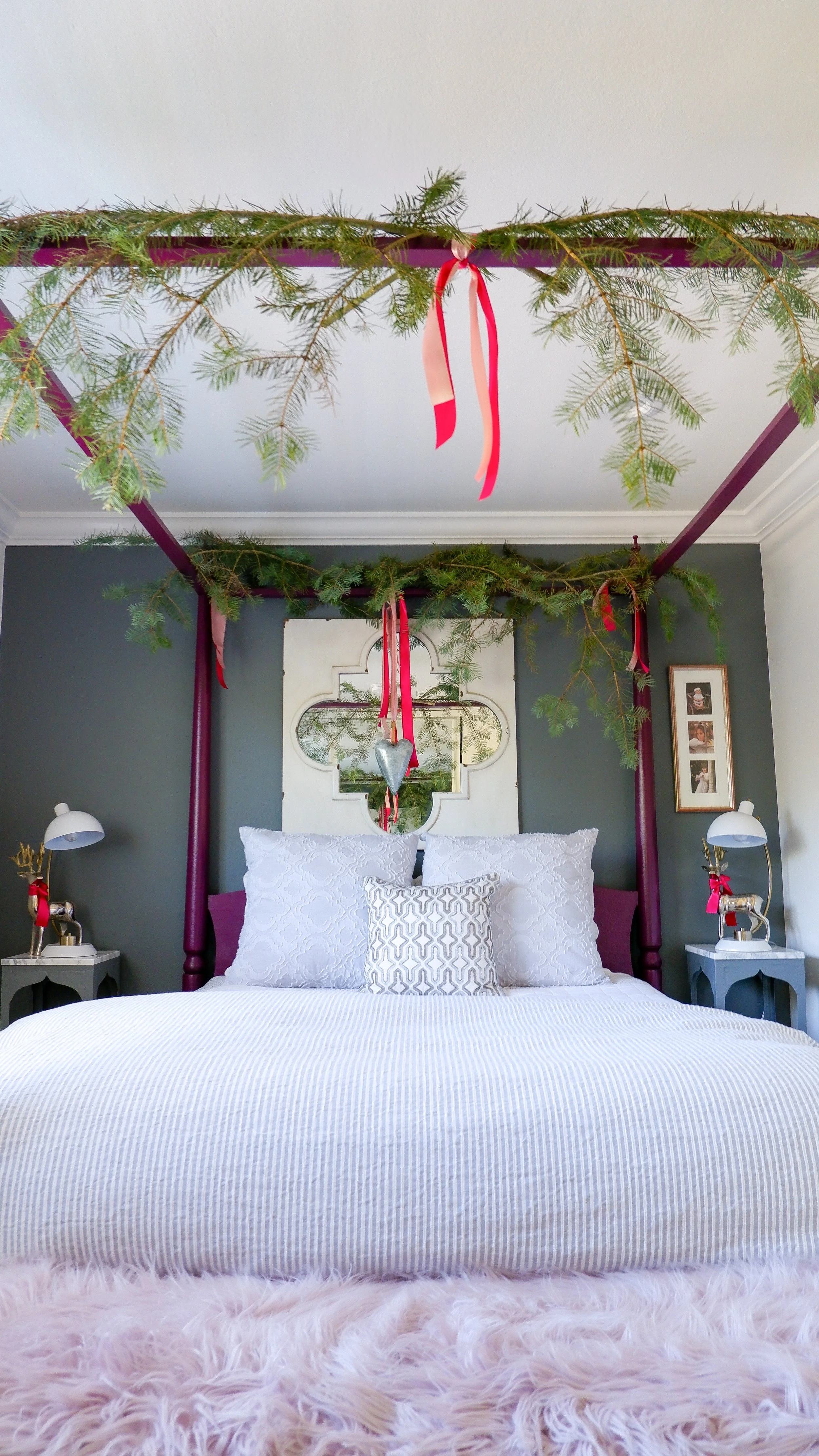 A bedroom with a four-poster bed decorated with pine garlands and red ribbons. The bed has white bedding and decorative pillows. There are side tables with lamps on both sides of the bed, and the wall behind features framed photographs and a decorative mirror.