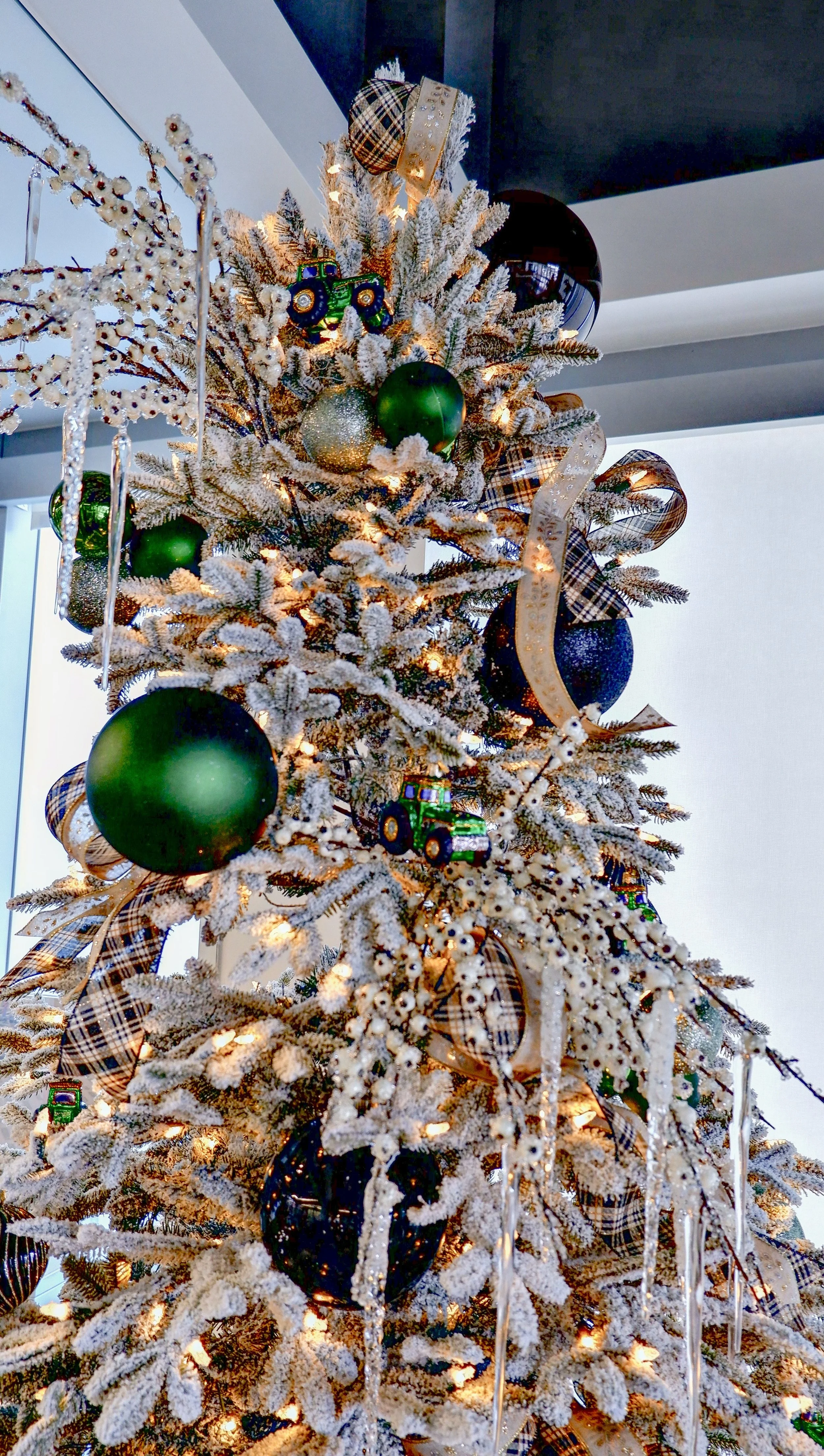 Christmas tree decorated with green and blue ornaments, plaid ribbon, tractor ornaments, and icicle decorations, lit with white lights.