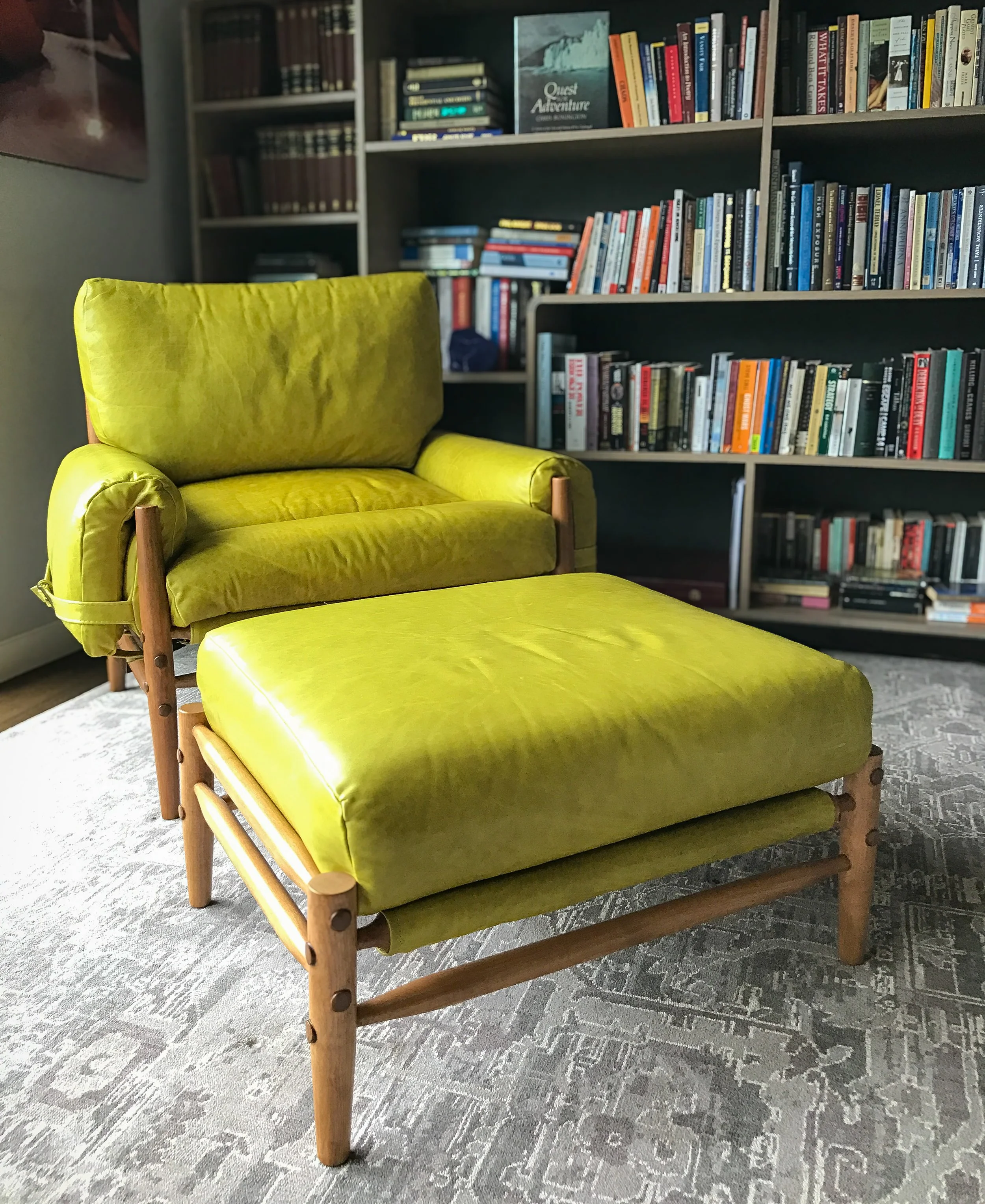 Lime green armchair with matching ottoman, wooden frame, in a room with bookshelves filled with books and a patterned gray carpet.