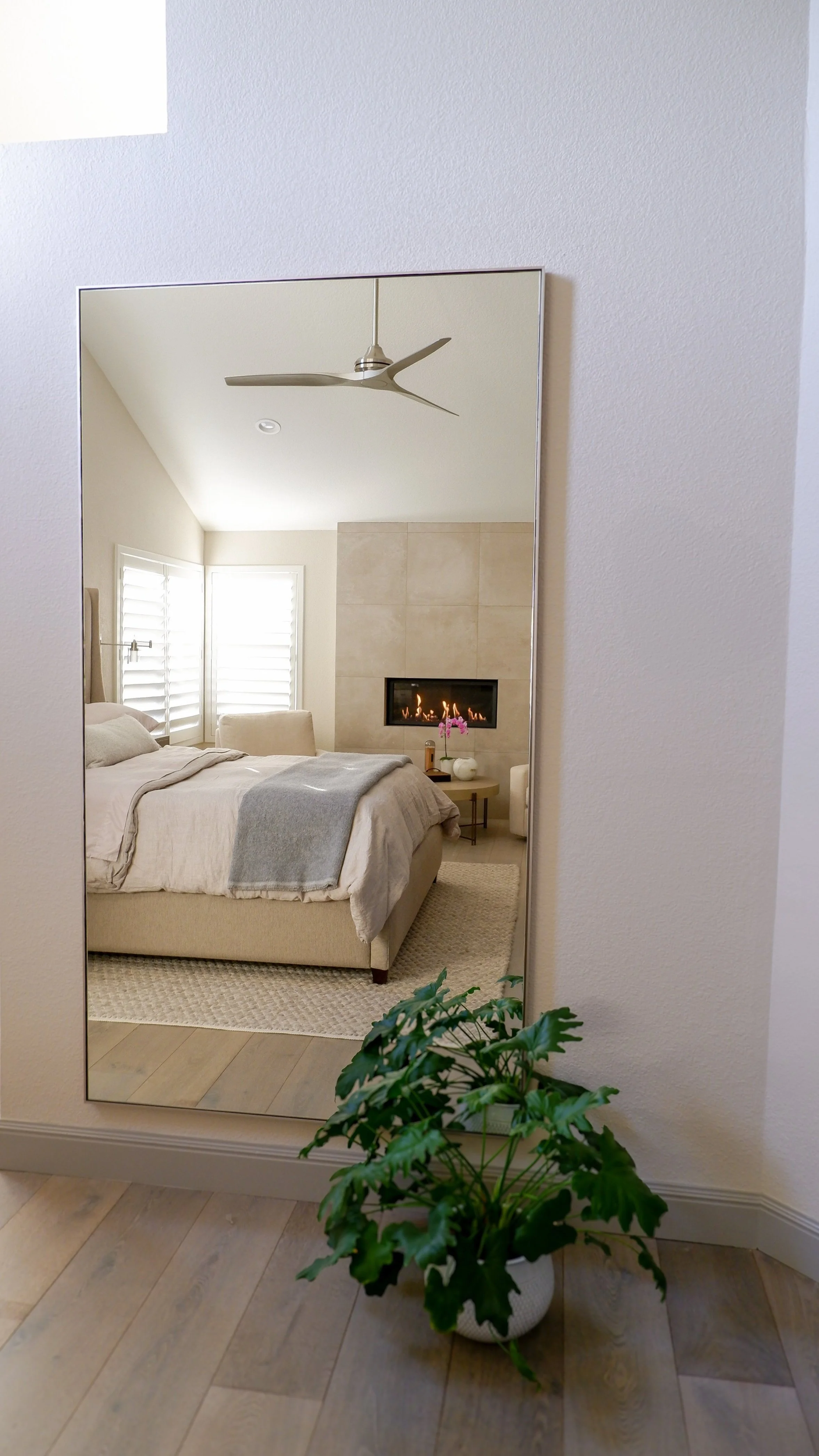 Reflection in mirror showing a bedroom with beige bedding, gray throw blanket, modern ceiling fan, fireplace, and a small plant on wooden flooring.