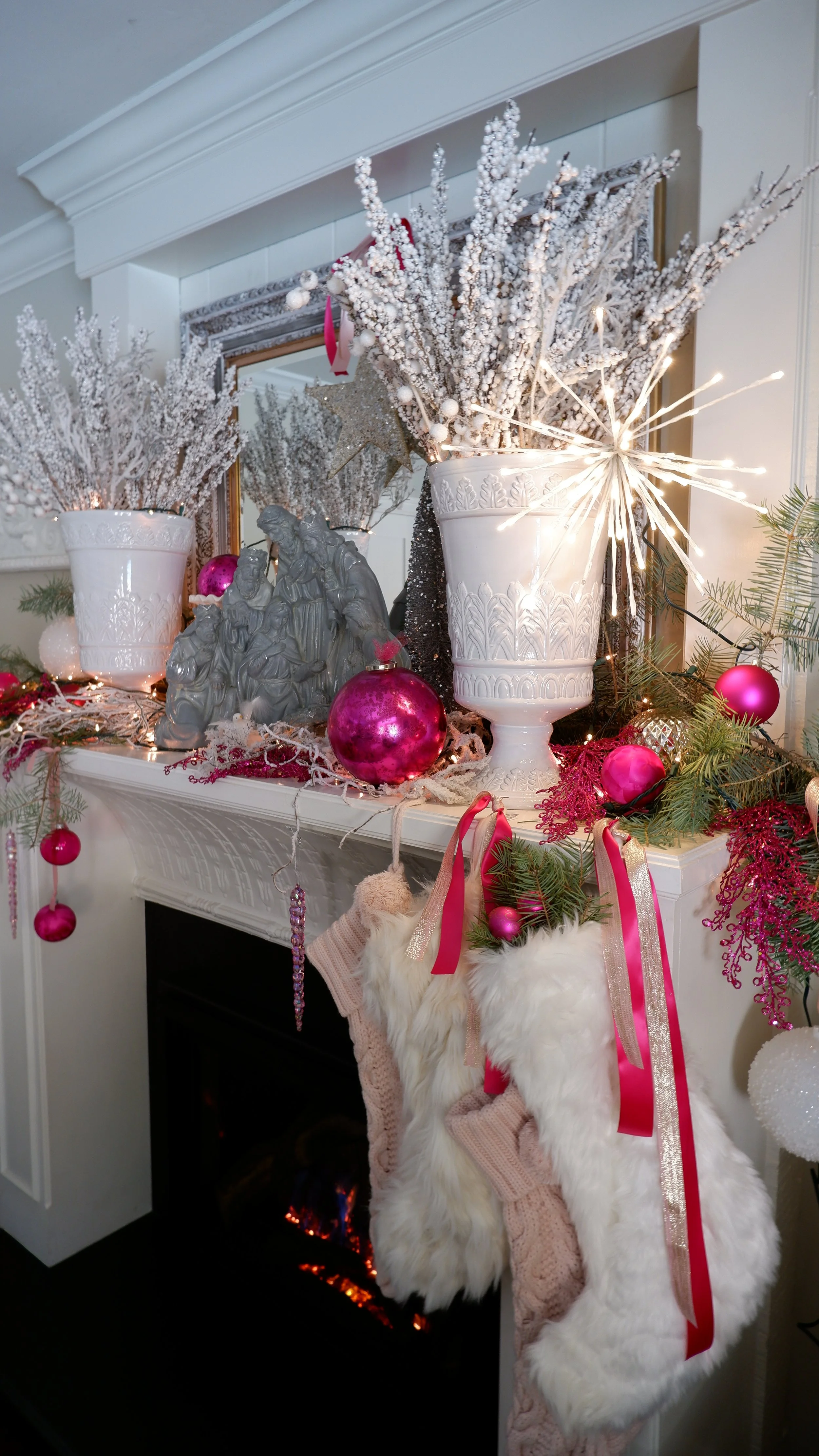 Christmas mantel decorated with white planters filled with frosted branches, pink ornaments, fairy lights, and a nativity scene. Pink and white stockings hang below.