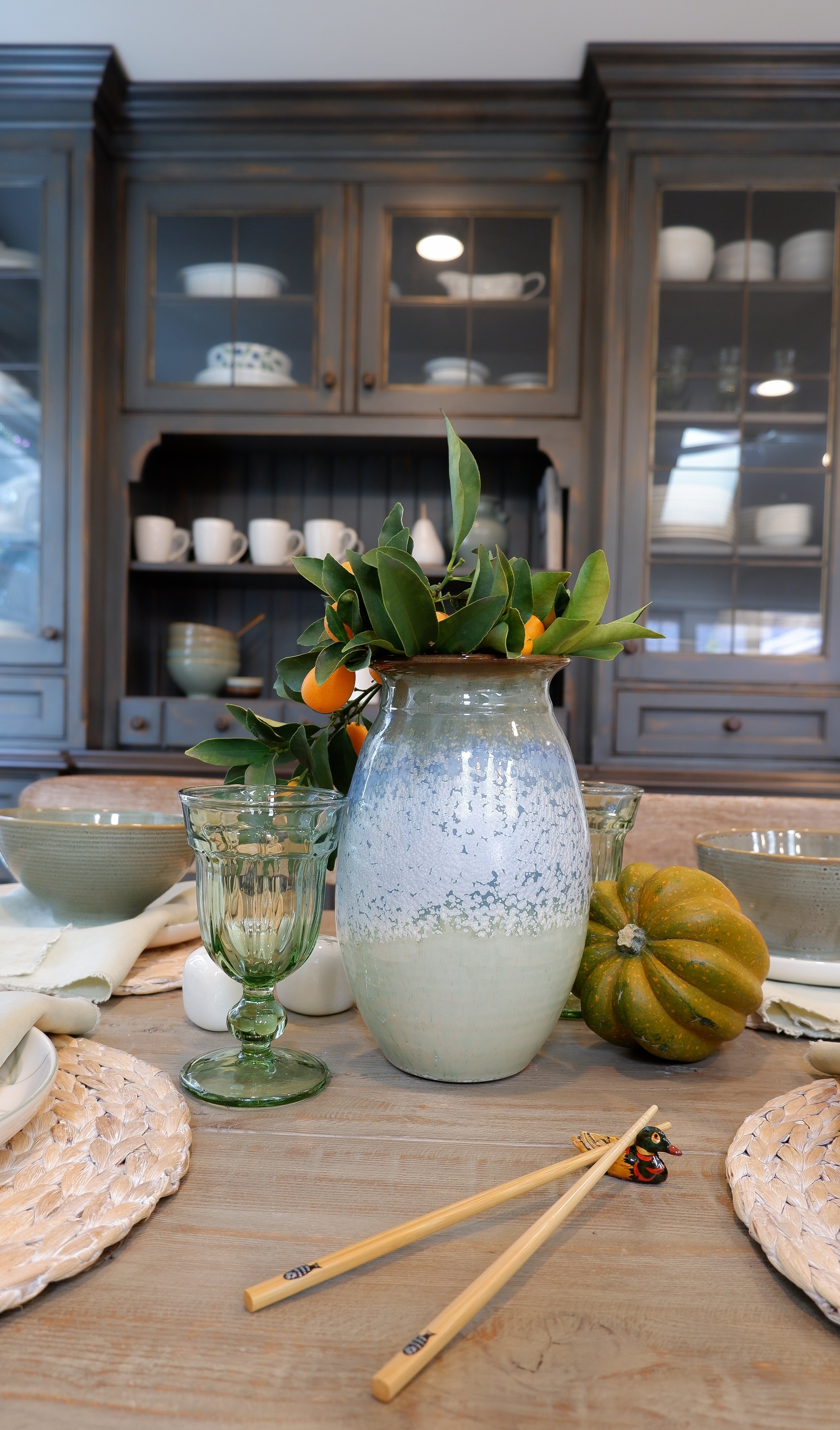 Table setting with ceramic vase containing branches, green glassware, small pumpkin, and chopsticks; gray hutch with dishes in background.