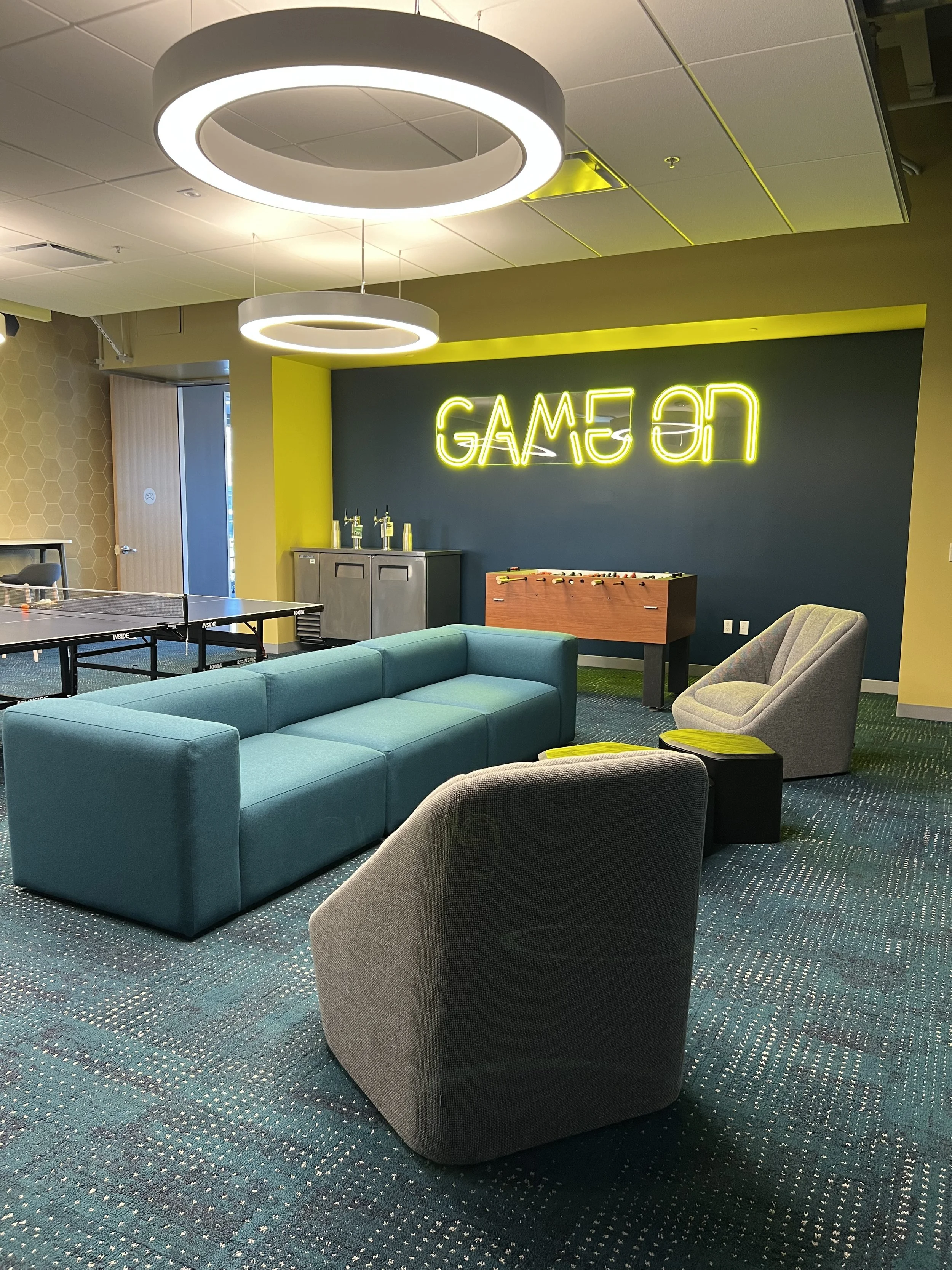 Modern game room with neon "Game On" sign, comfortable seating, foosball table, and ping pong table.