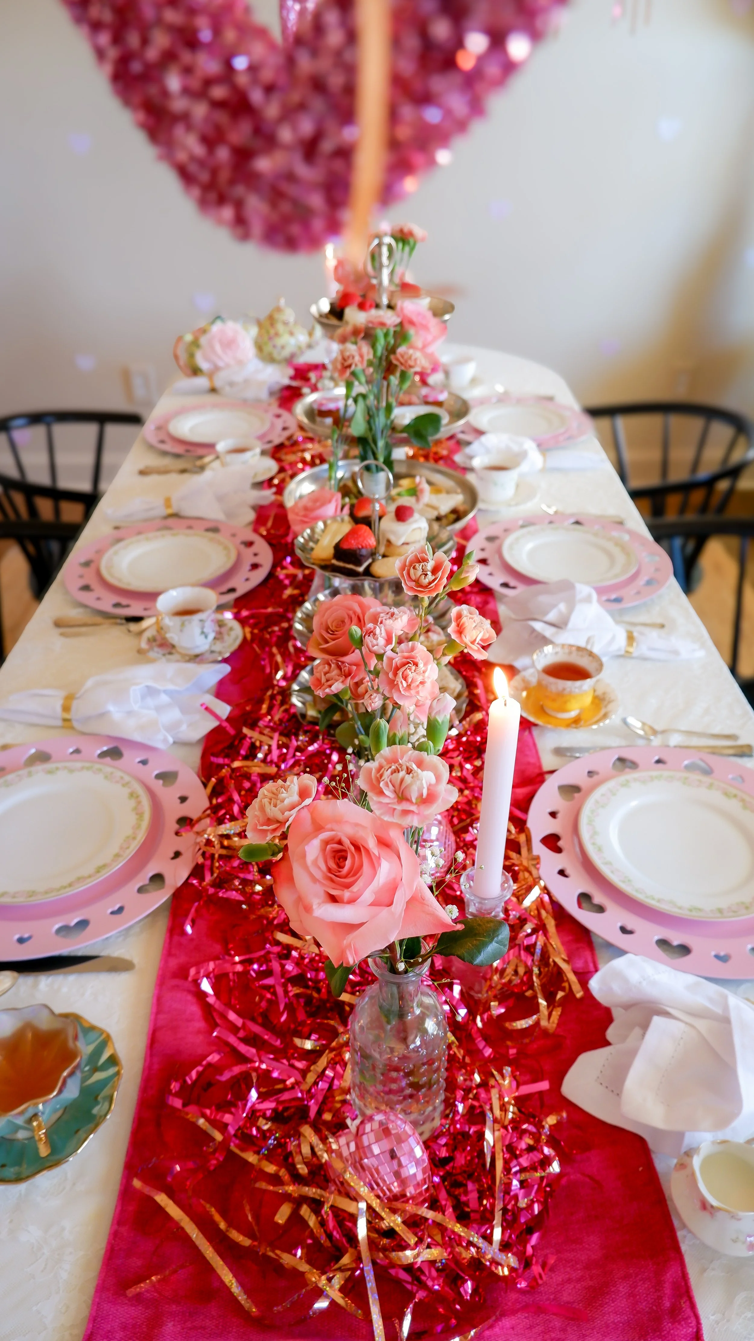Decorated table set for a Valentine’s Day event, featuring a long pink runner with roses and carnations, heart-themed plates, tea cups, and a heart-shaped pink balloon overhead.