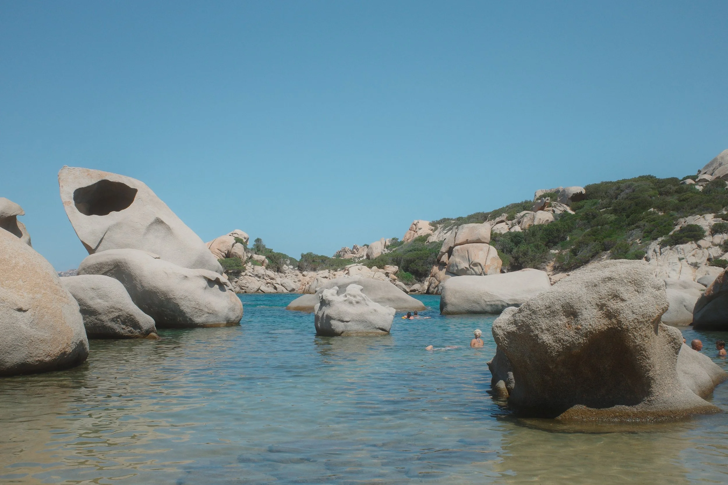 People swimming in a rocky, coastal area with large granite boulders and clear blue water under a bright blue sky.
