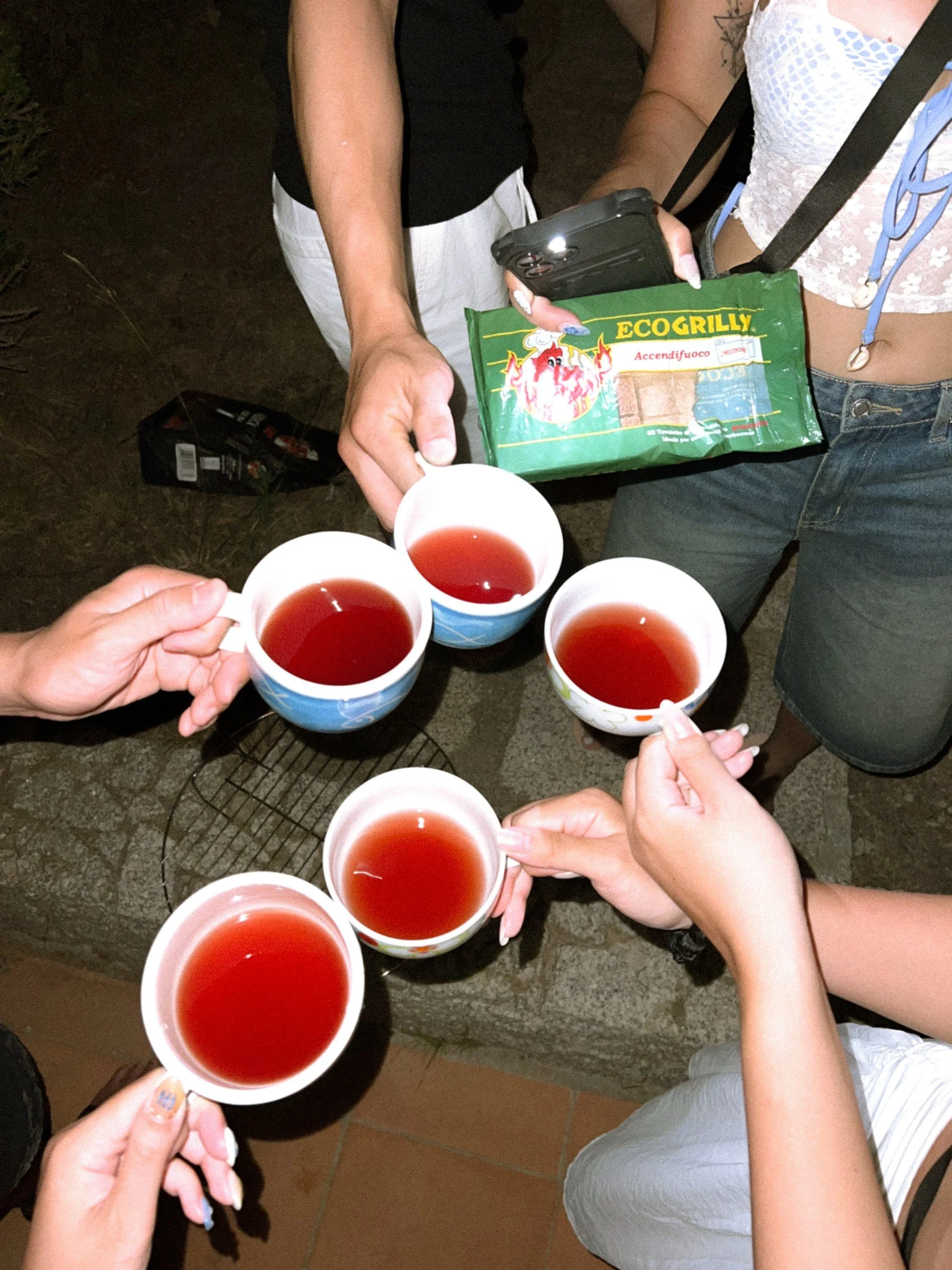 Group of people holding white cups of tea, standing around a stone surface at night.