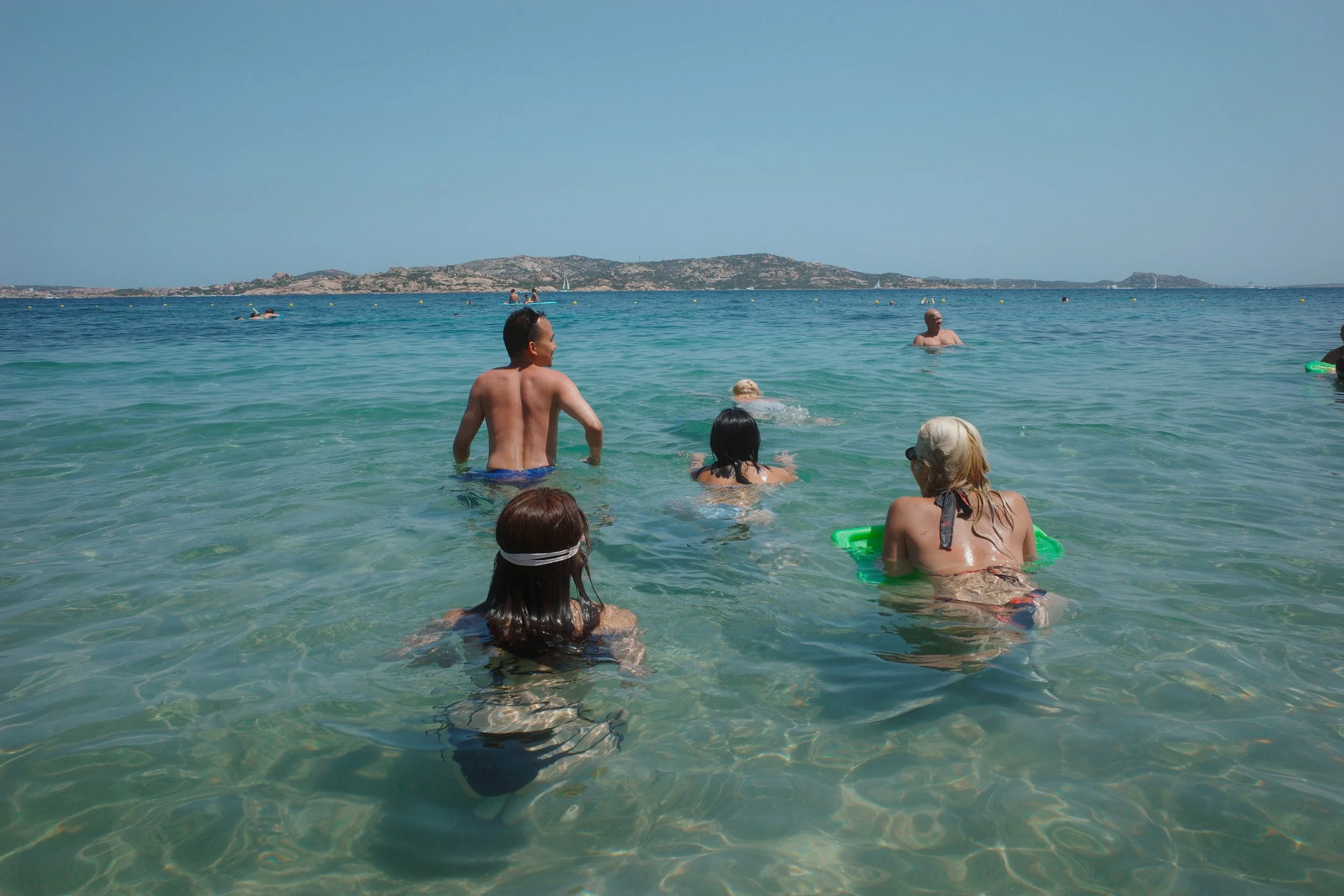 People swimming and relaxing in clear ocean water near a beach with a landmass in the distance on a sunny day.