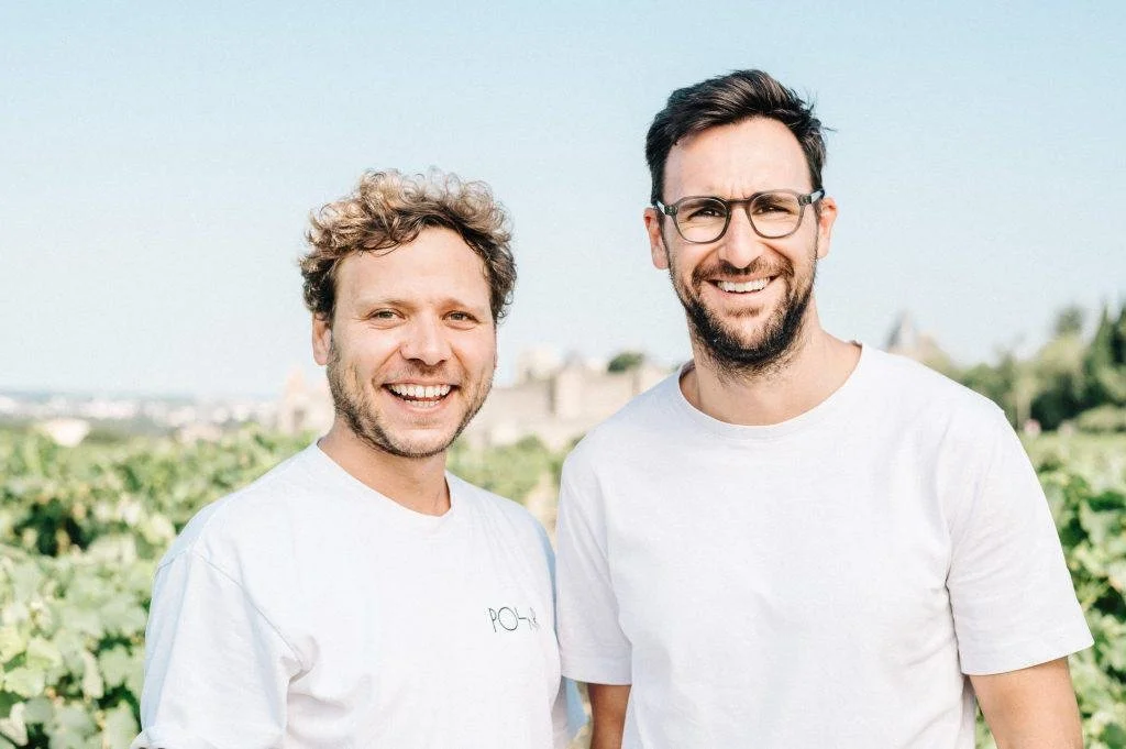Two smiling men standing outdoors in a vineyard, wearing white t-shirts, with a bright blue sky in the background.