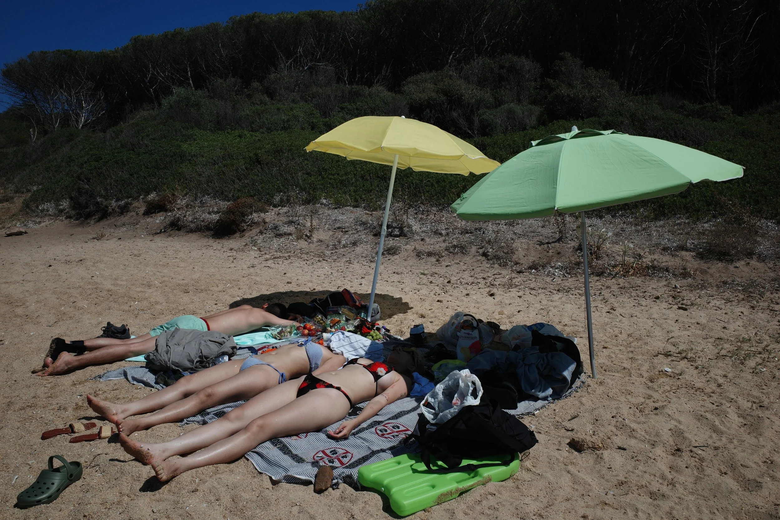 Three people lying on beach towels and sunscreen bottles on the sand under yellow and green beach umbrellas, with beach bags and shoes nearby, and a background of bushes and trees.