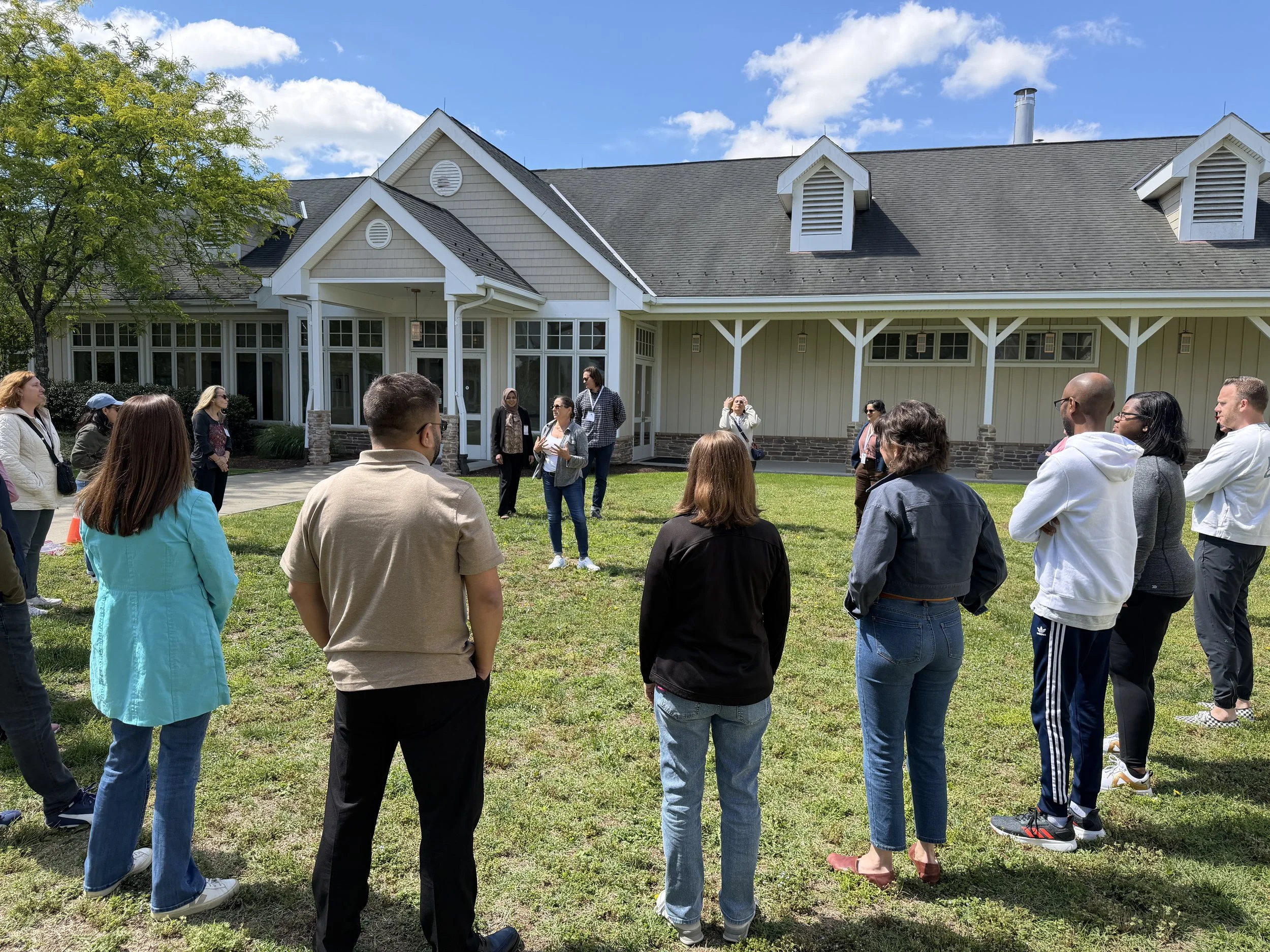 Photo of Debbie Fink in the center of a circle of ~25 diverse participants, instructing them in the "why" of what awaits. They are all outdoors on grass, with a building in the background and blue skies.