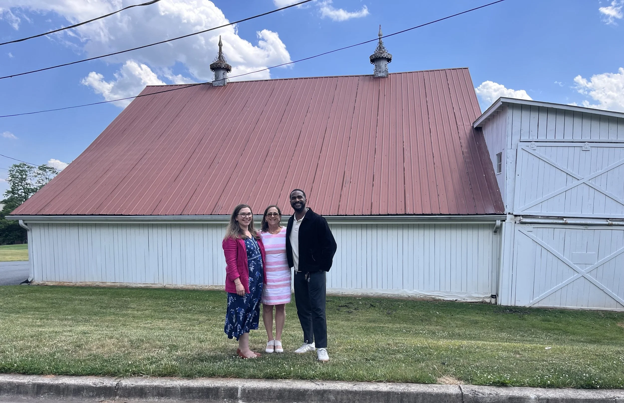 Debbie standing in between two mentees, outdoors on a blue-sky day, with a red and white barn behind them. They are arm in arm, smiling at camera.