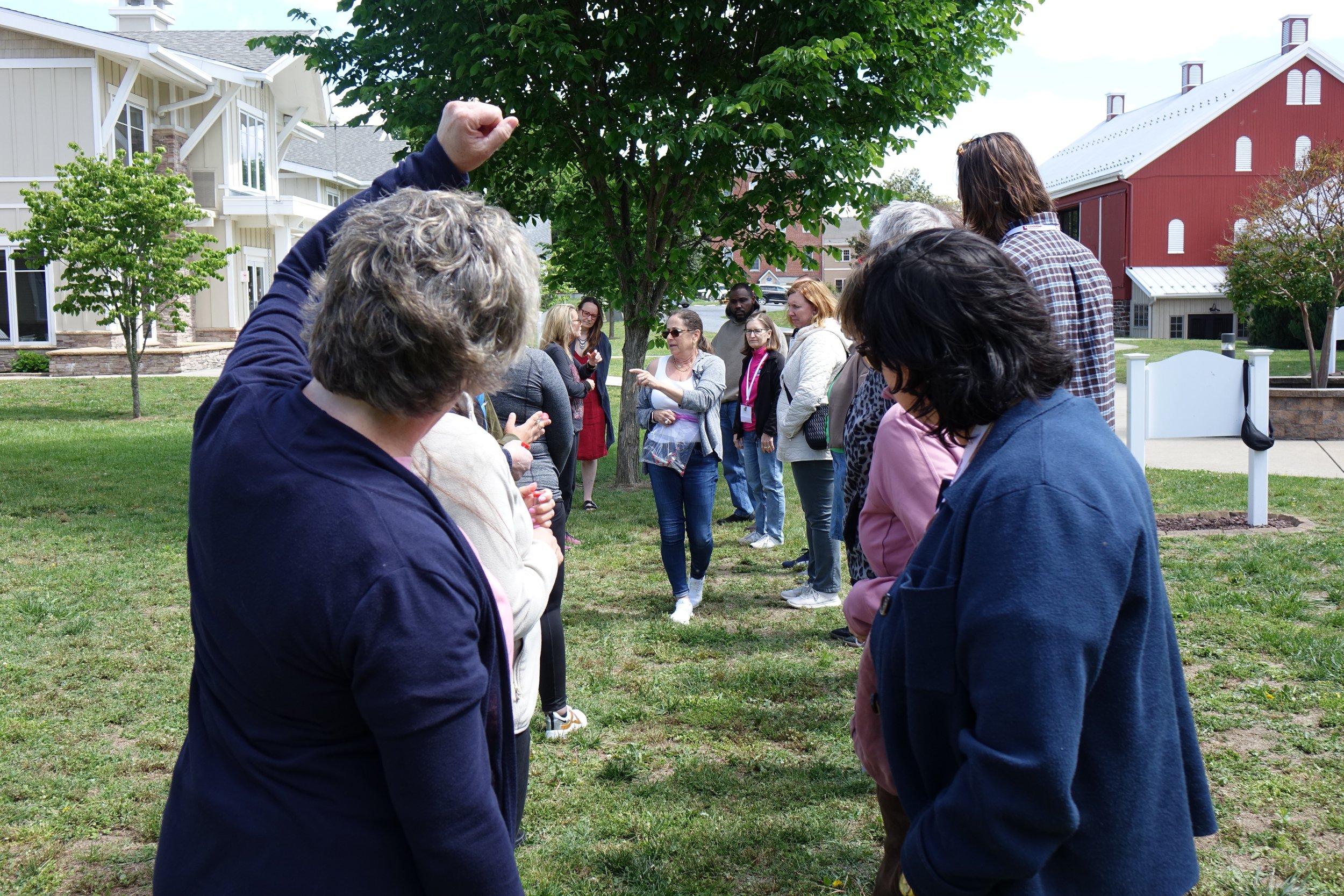 Two rows of diverse participants stand outside, with Debbie Fink in the middle, providing directives for this exercise. Debbie holds a bag of one-minute timers. A tree, barn, and building are in the background.