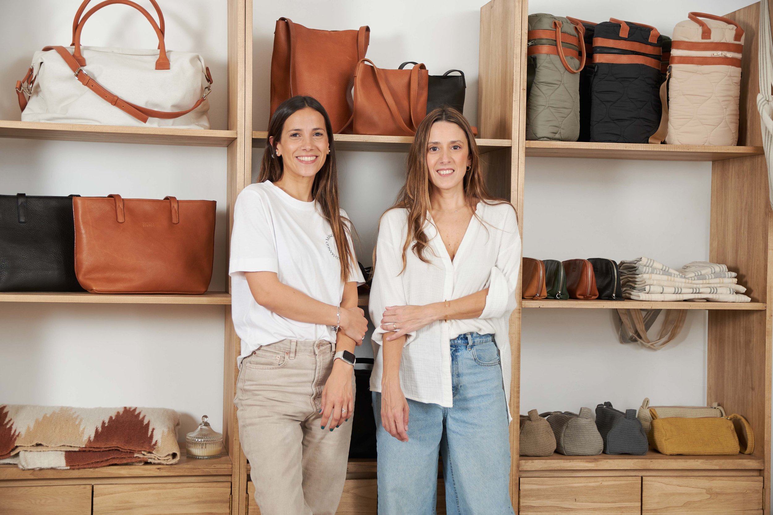 Dos mujeres sonrientes en una tienda de bolsos, rodeadas de estanterías con bolsos y accesorios de diferentes colores y tamaños.
