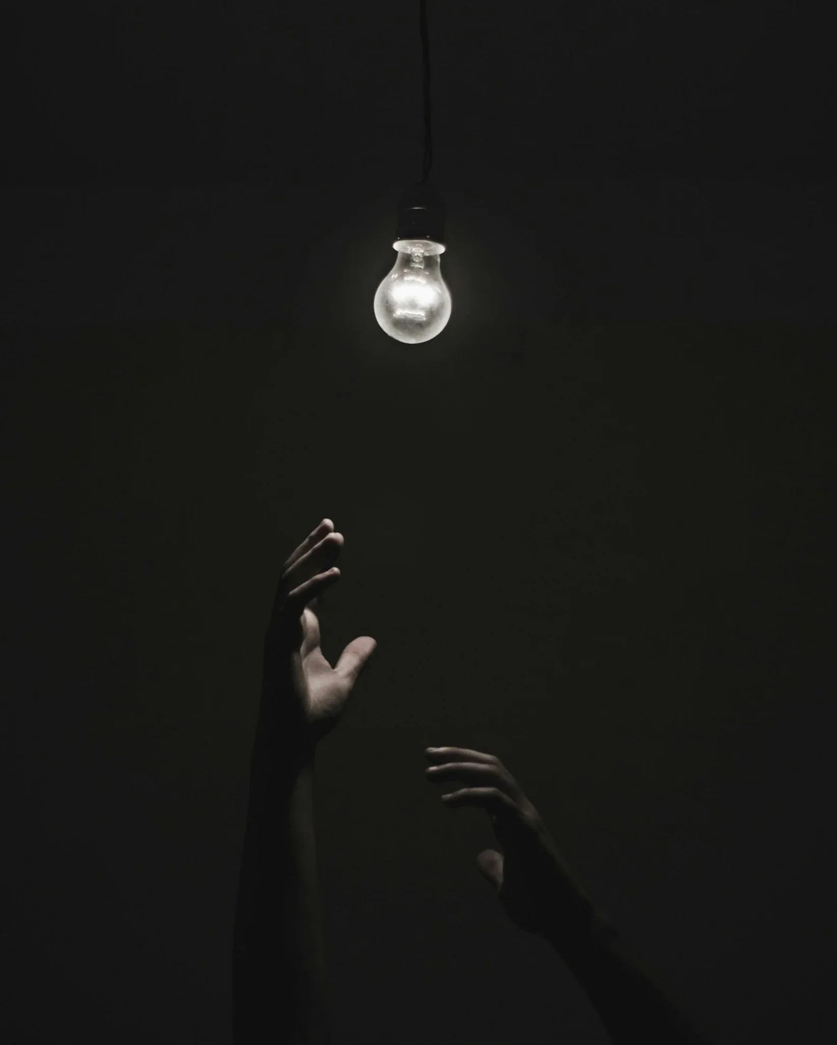 Dark image of a incandescent light bulb hanging from the ceiling with a person's hands reaching upward beneath it.