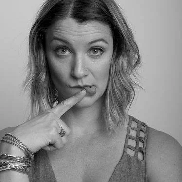 Black and white photo of a woman with wavy hair, one finger is pressing her lower lip to the side and she has one eyebrow raised