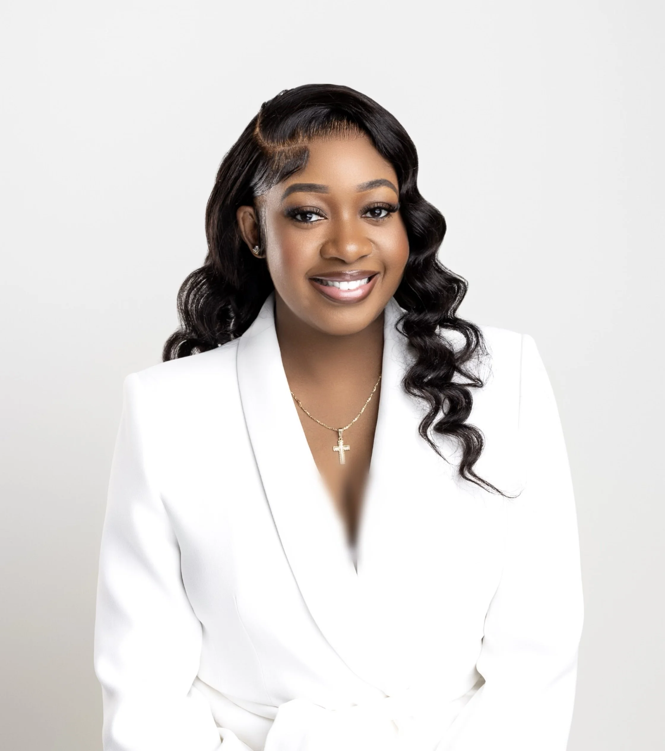 Person smiling in a white suit with curly hair and a cross necklace, against a light background.