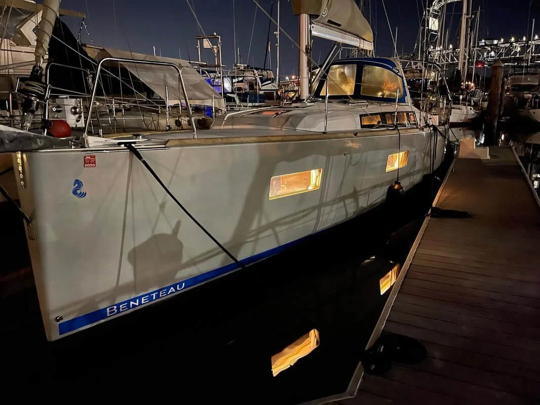 A Beneteau sailboat docked at a marina at night, with its cabin lights on, casting reflections on the water. Other moored boats and a bridge are visible in the background.