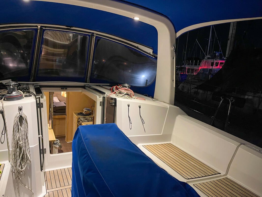 Interior of a yacht at night, showing the cockpit with a blue cover and a view into the cabin with wooden furnishings. Outdoor lighting highlights nearby boats.