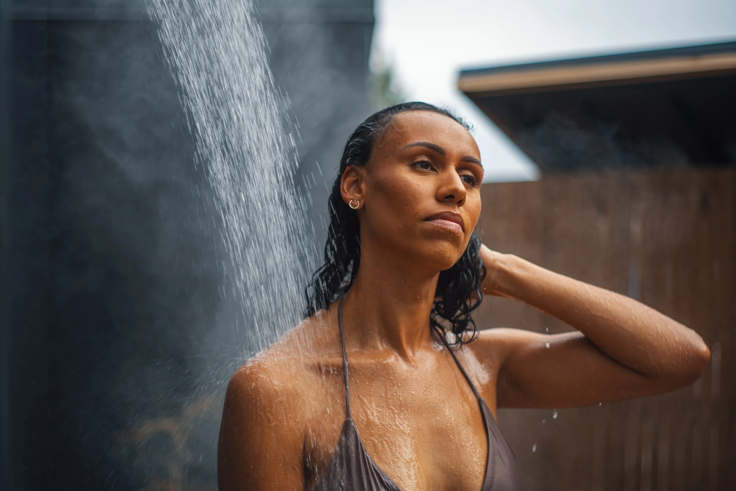 Woman with wet hair and a brown swimsuit standing under outdoor showerhead