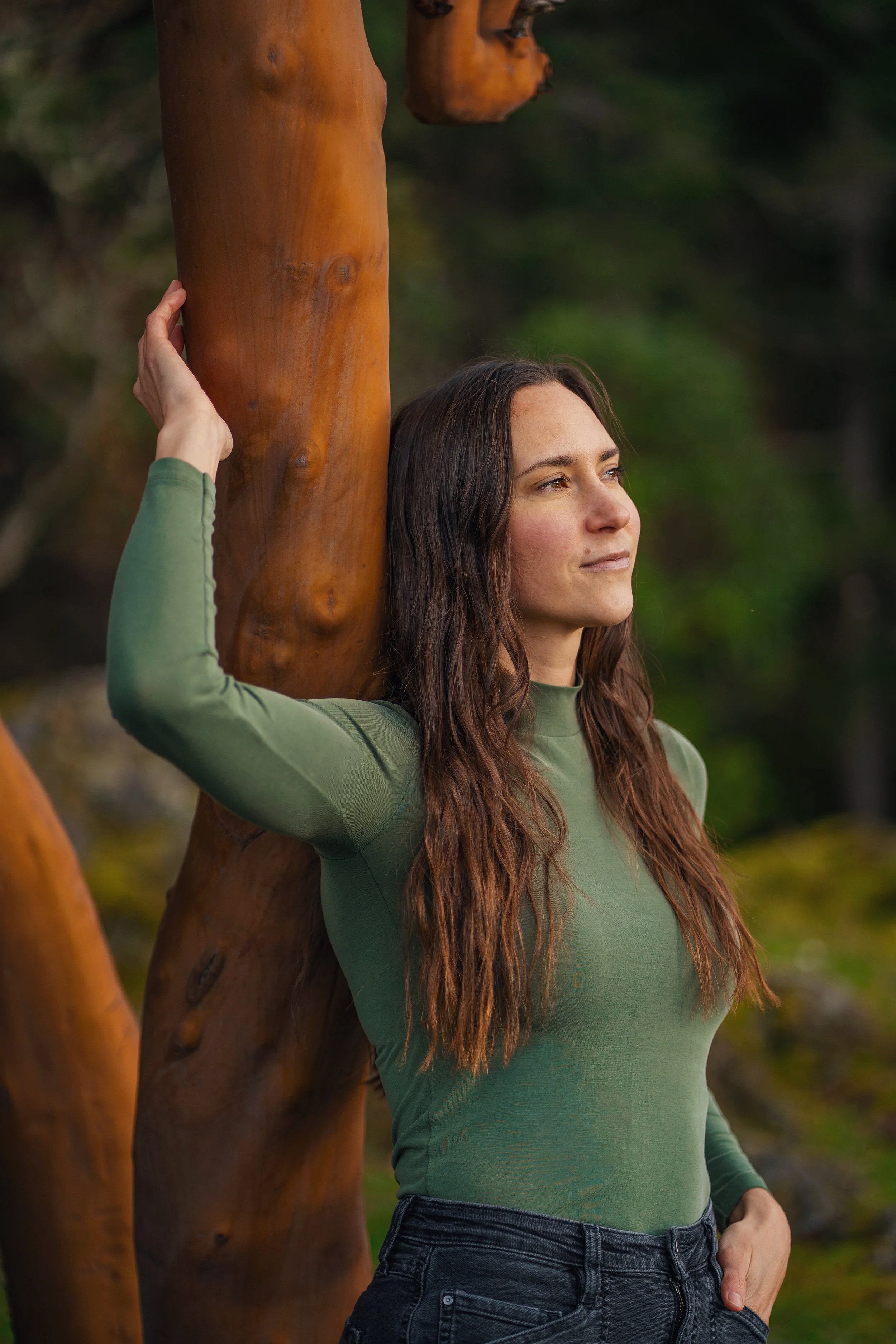 A woman with long brown hair wearing a green long-sleeved shirt and black jeans, standing next to a large wooden sculpture of a horse's body, outdoors with a blurred background of trees.