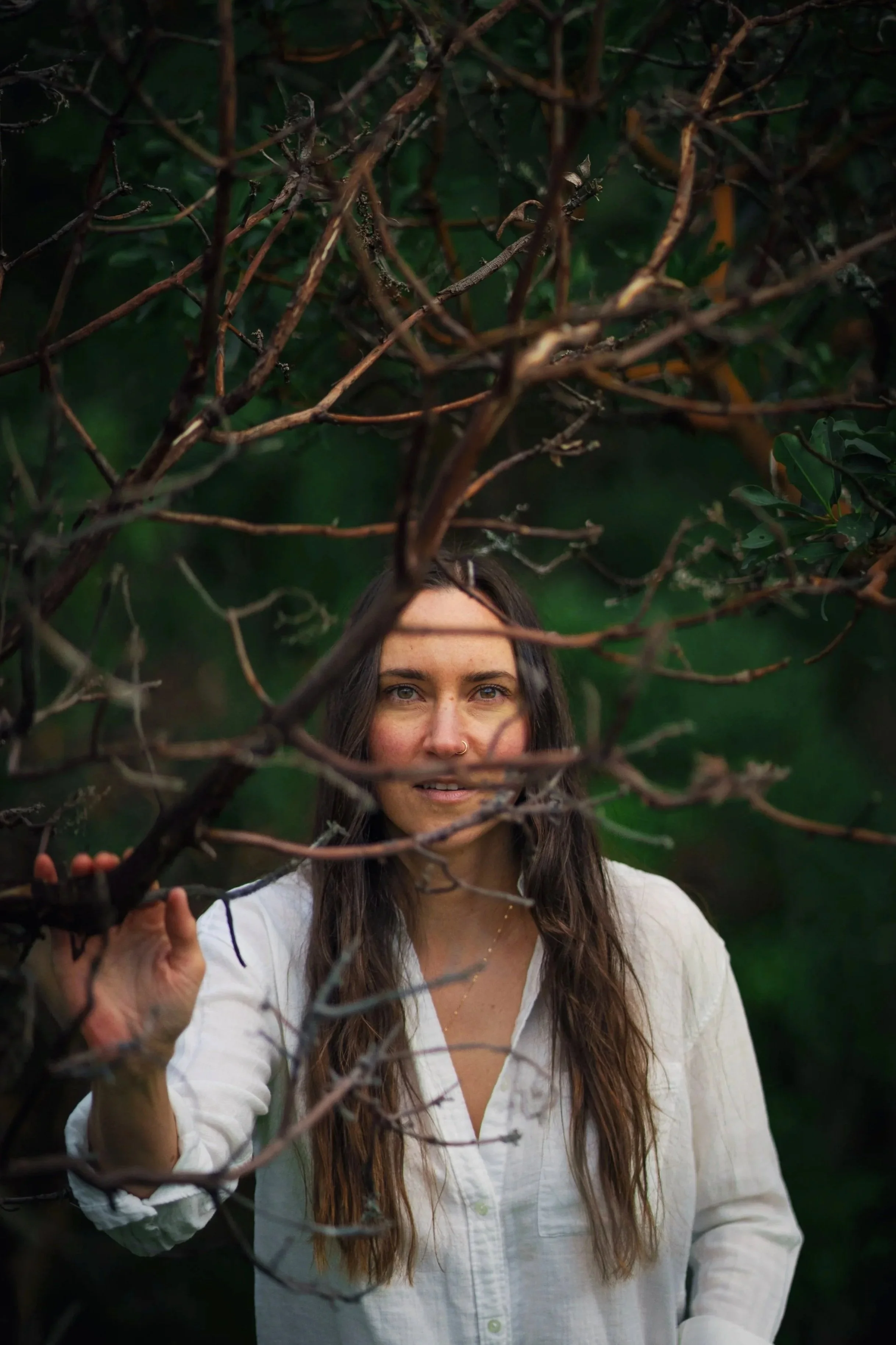 A woman with long brown hair and a white shirt looking through bare tree branches.