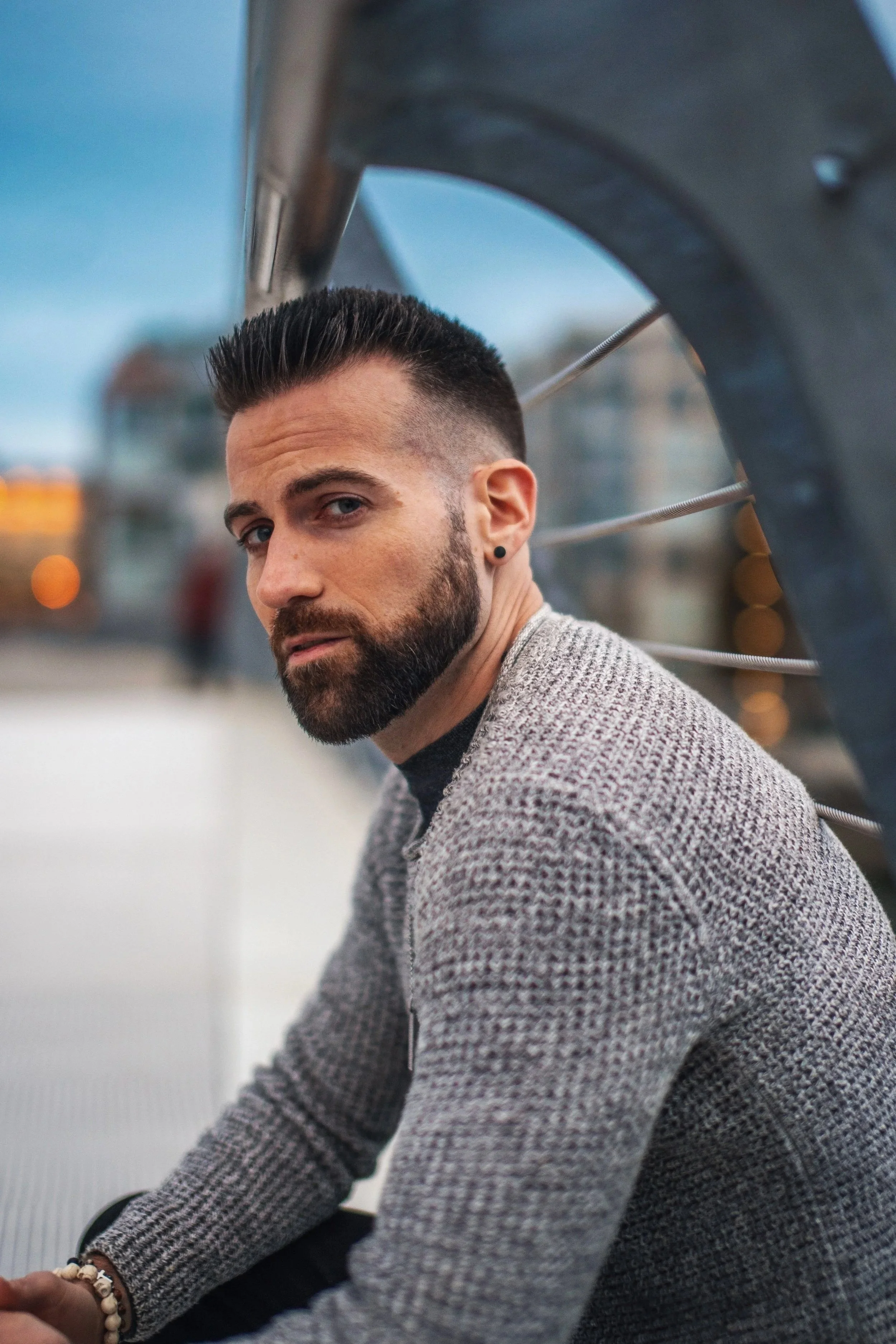 A man with a beard and short dark hair, wearing a gray knit sweater, looks at the camera while sitting outdoors near a railing.