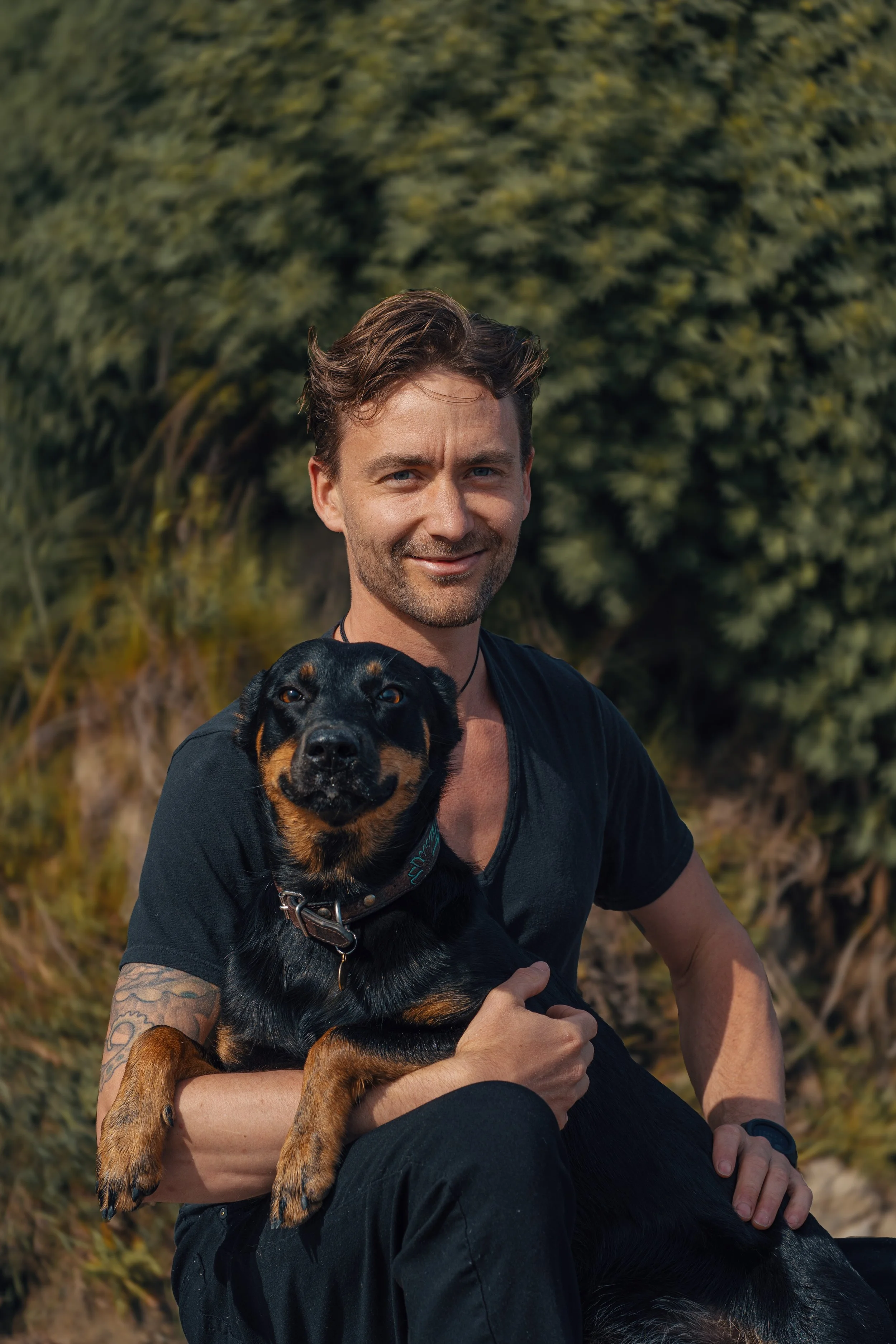 A man with brown hair, blue eyes, and a beard sitting outdoors with his dog in front of green trees. The man is smiling slightly, wearing a black shirt, and holding the black and brown dog, which is looking at the camera.