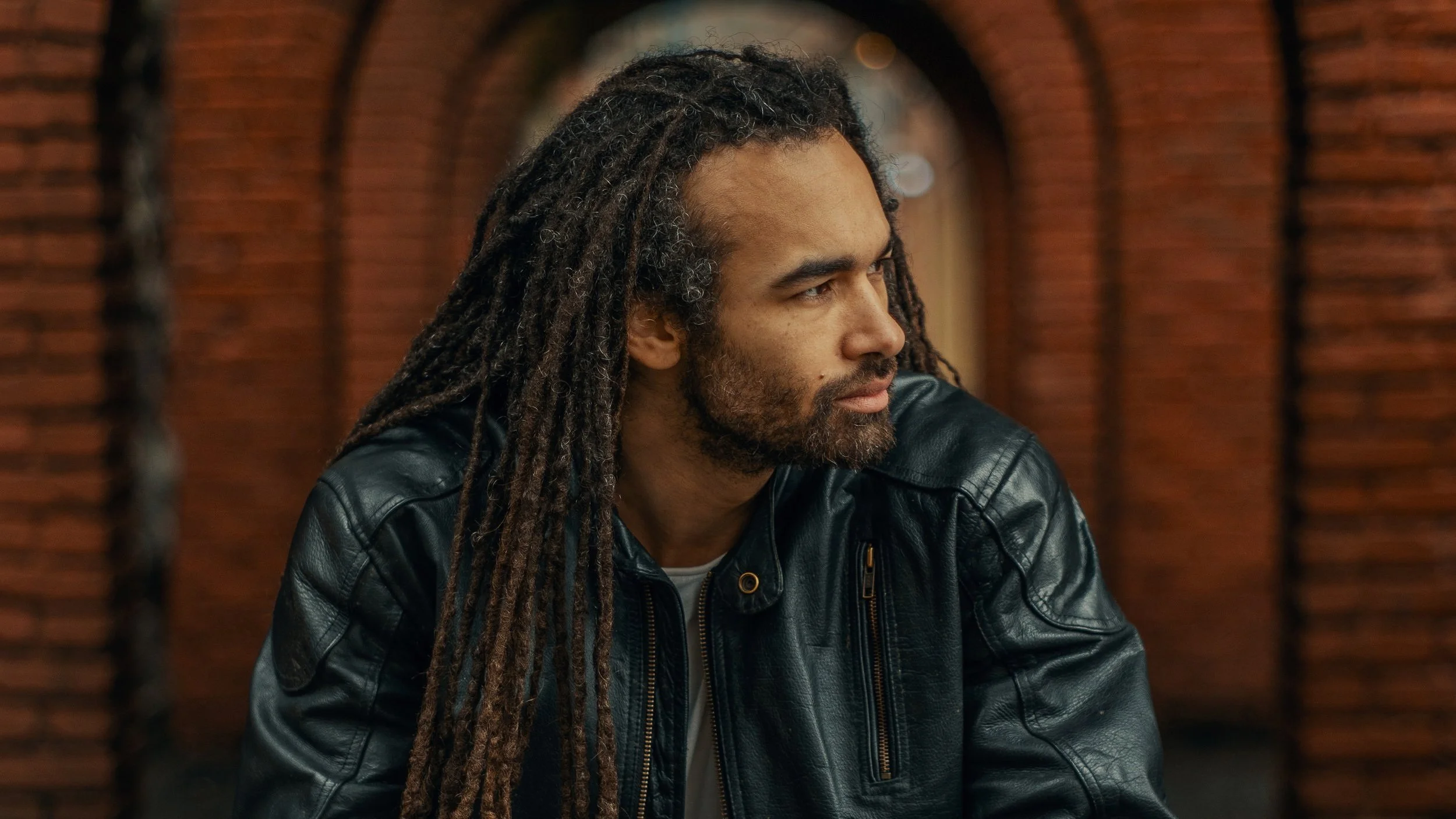 A man with dreadlocks and a beard, wearing a black leather jacket, sitting outdoors against a background of brown brick arches.