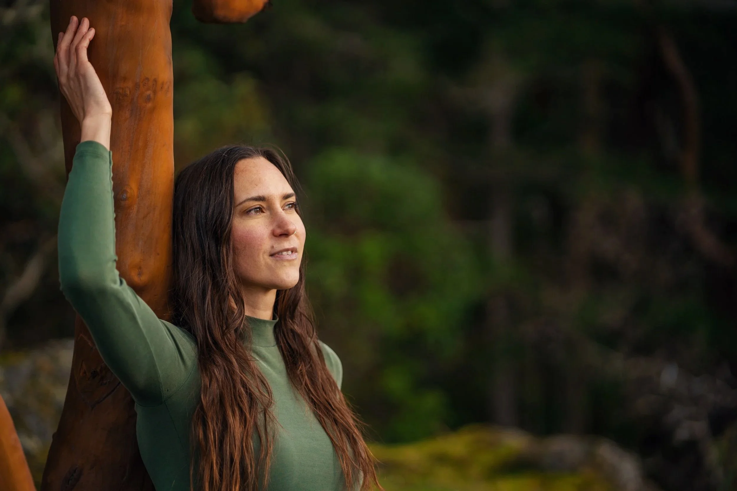 a woman with long brown hair wearing a green long-sleeve shirt, standing outdoors leaning against a tree trunk, with a blurred green forest background